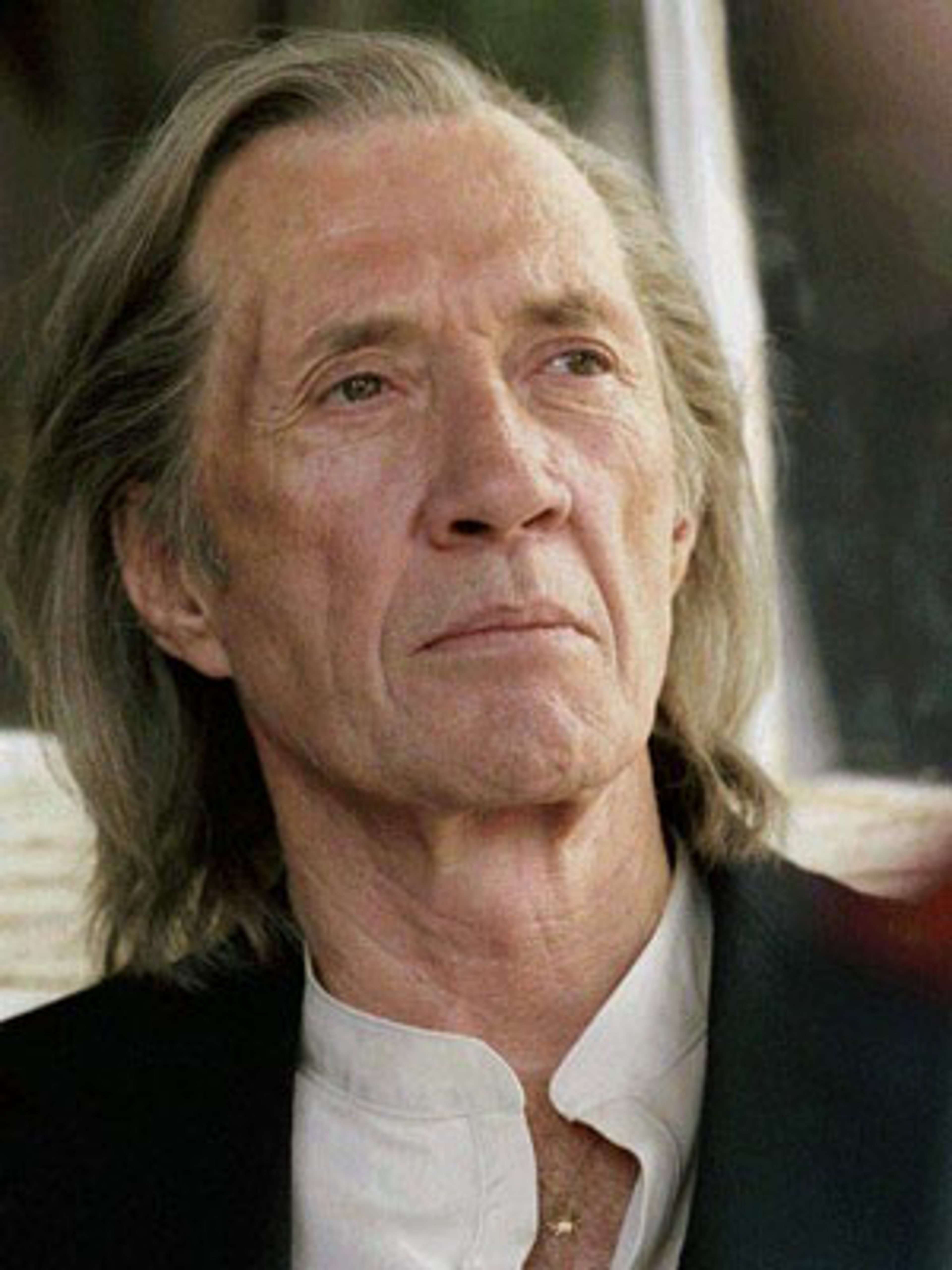 An older man with long gray hair and a beard, wearing a white shirt and black suit jacket, with a serious expression on his face.