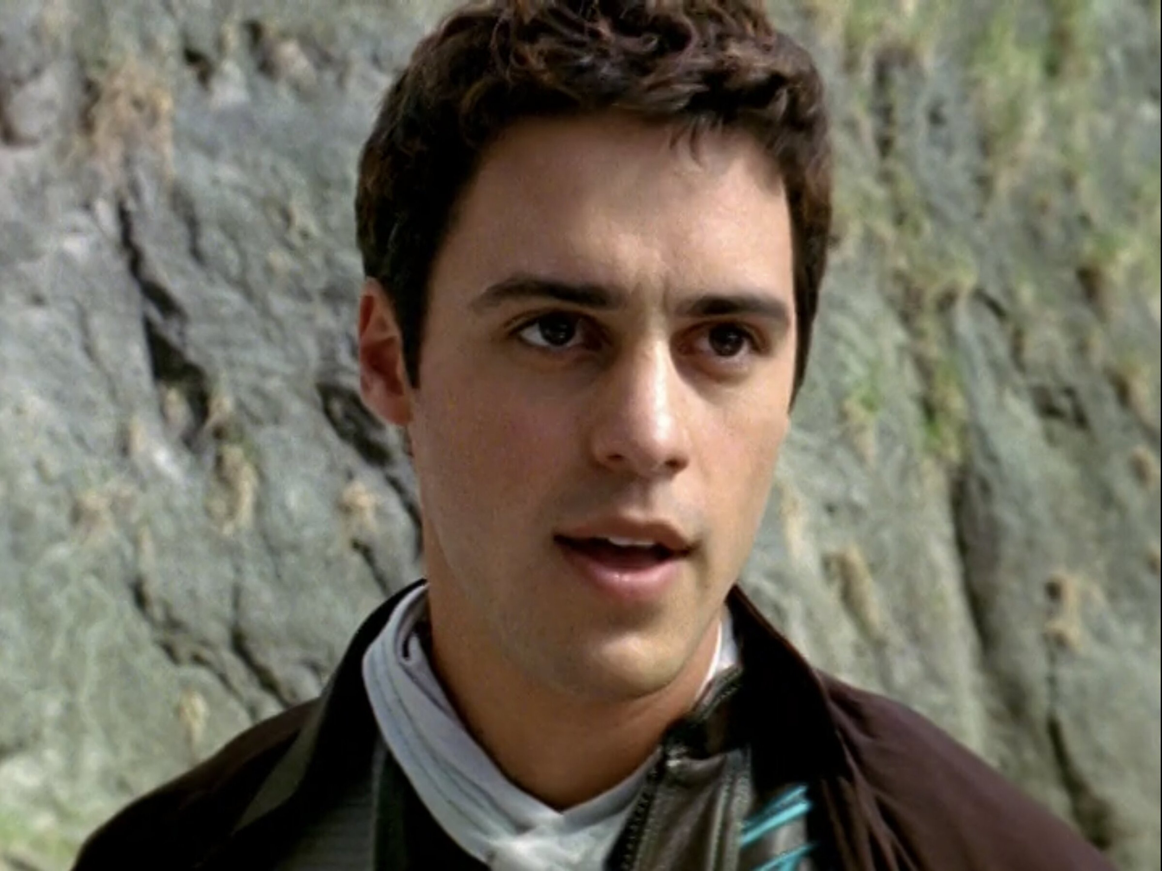 A young man with short dark hair wearing a green jacket, standing outdoors with a rocky background.