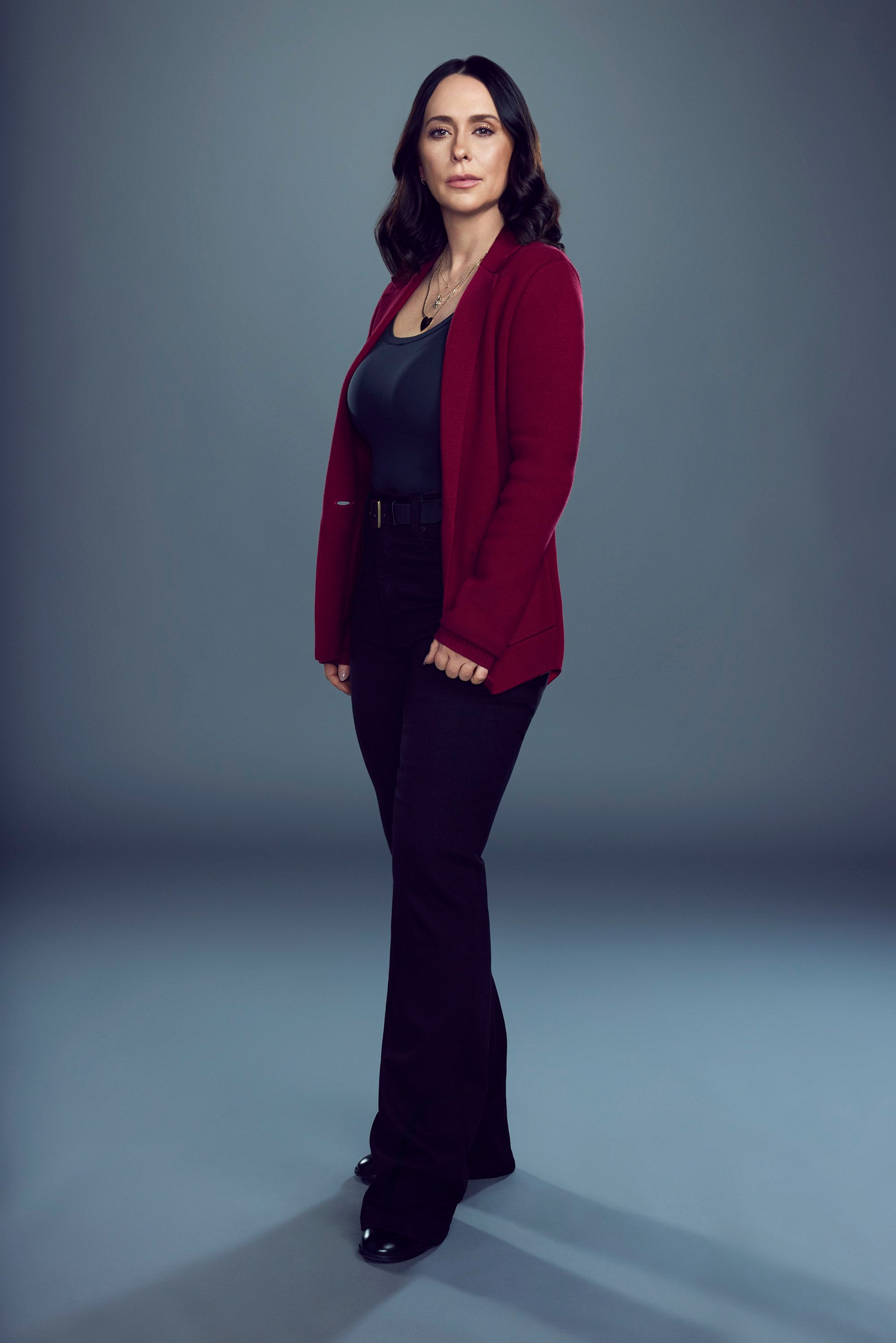 A woman with dark hair wearing a red cardigan and black dress, standing in a studio setting with a serious expression.