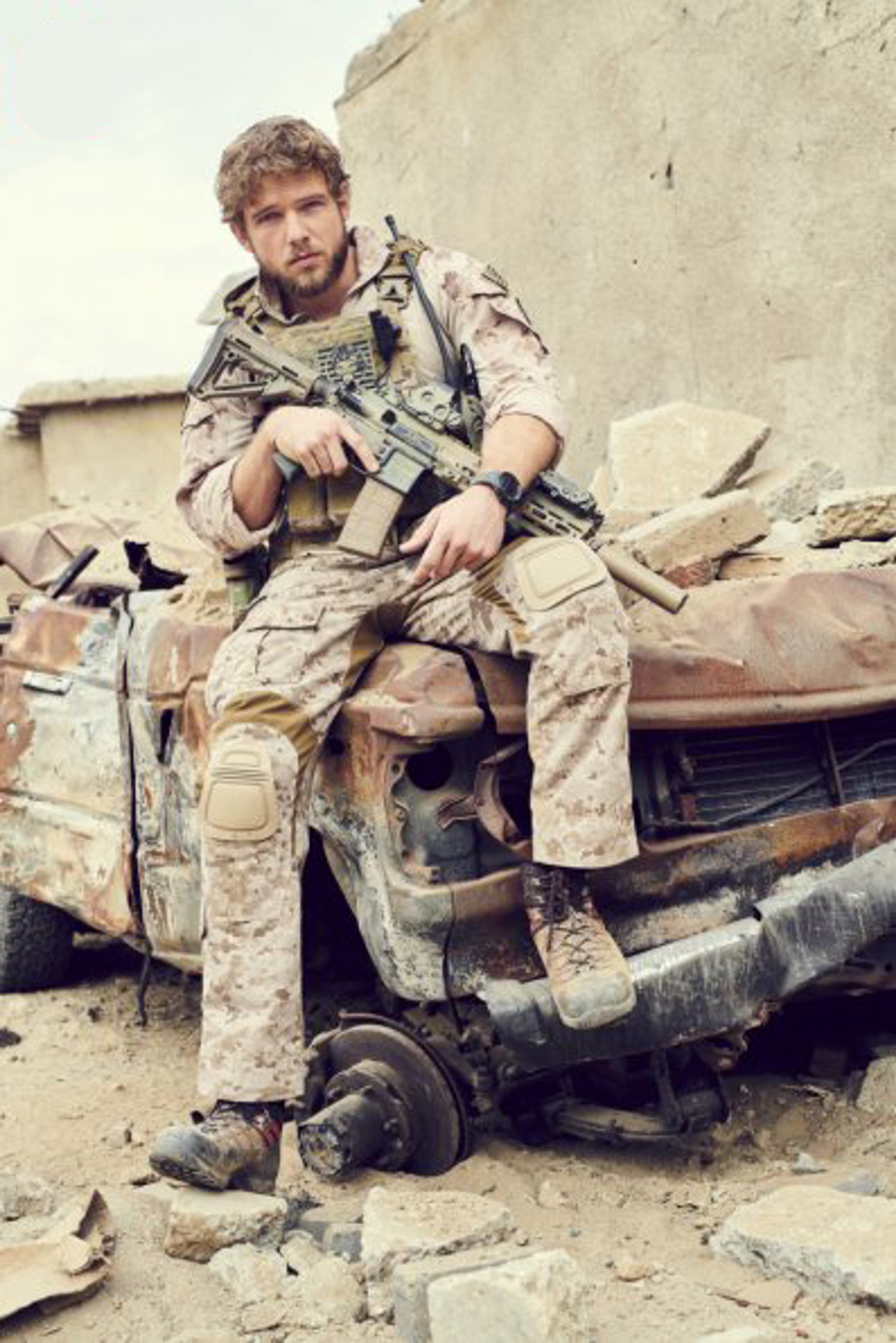 A man in military gear sitting on a damaged vehicle in a desert or rocky setting