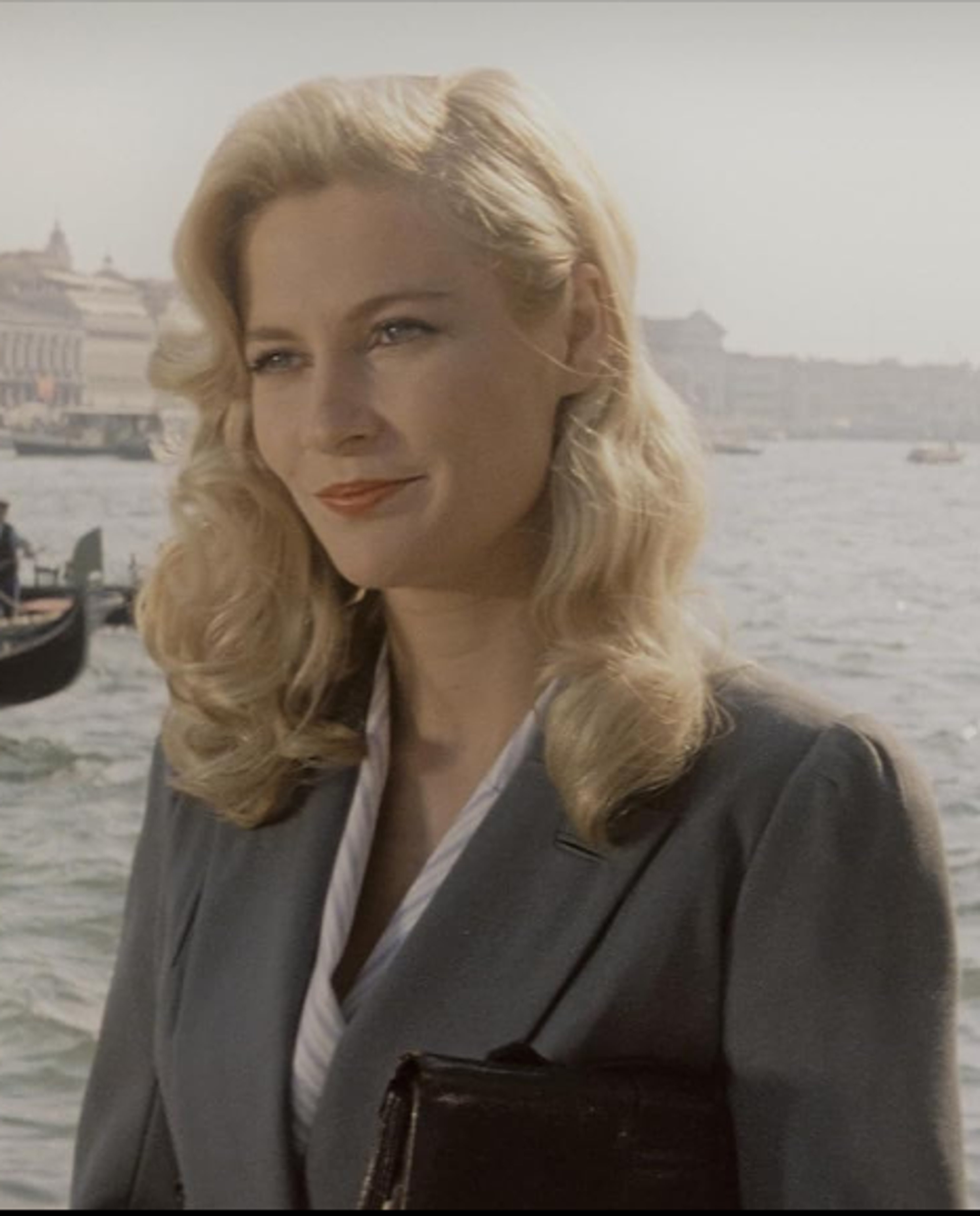 A woman with blonde hair wearing a gray coat standing in front of a body of water