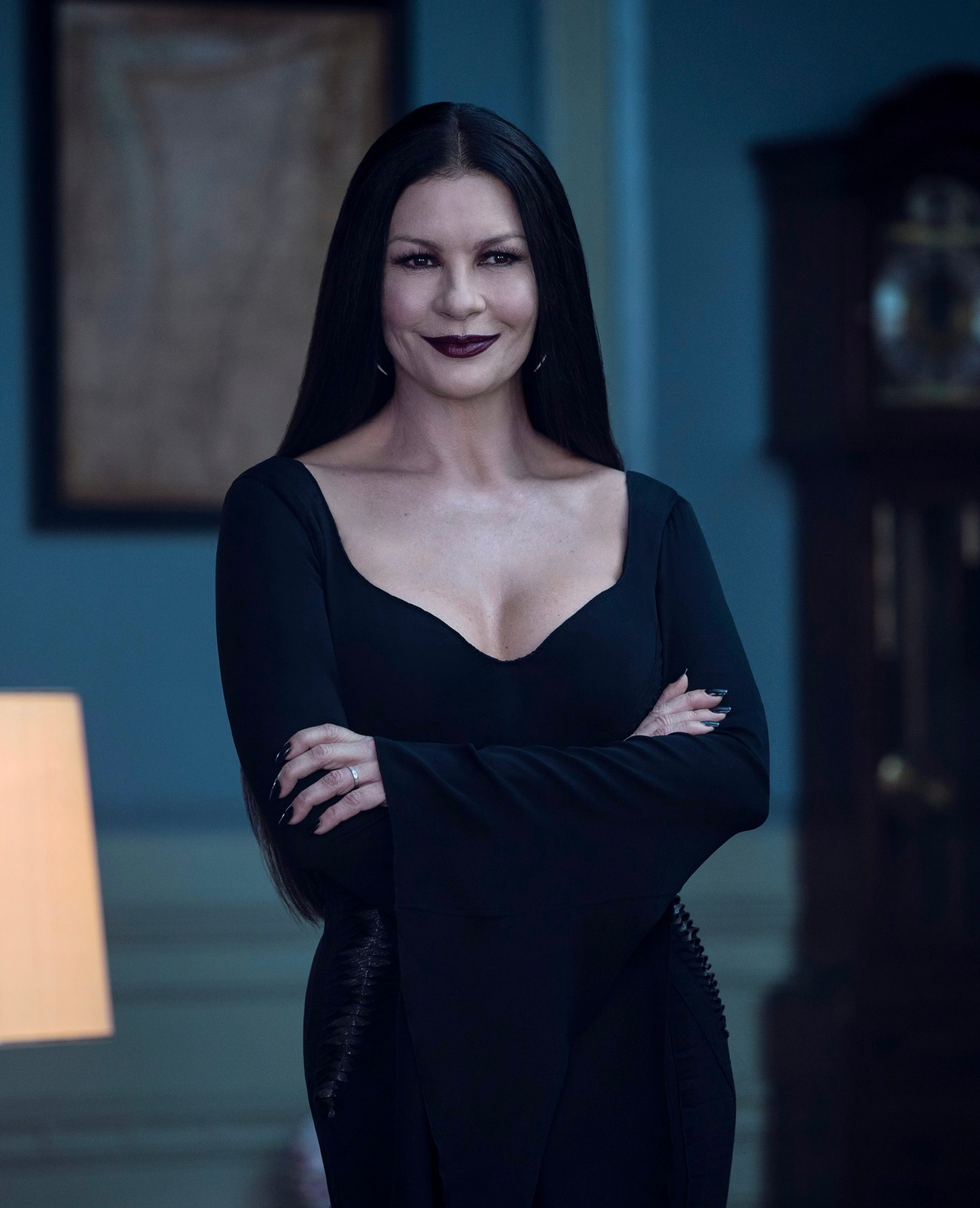 A woman with pale skin, dark hair, and a black dress standing in a room with a serious expression.