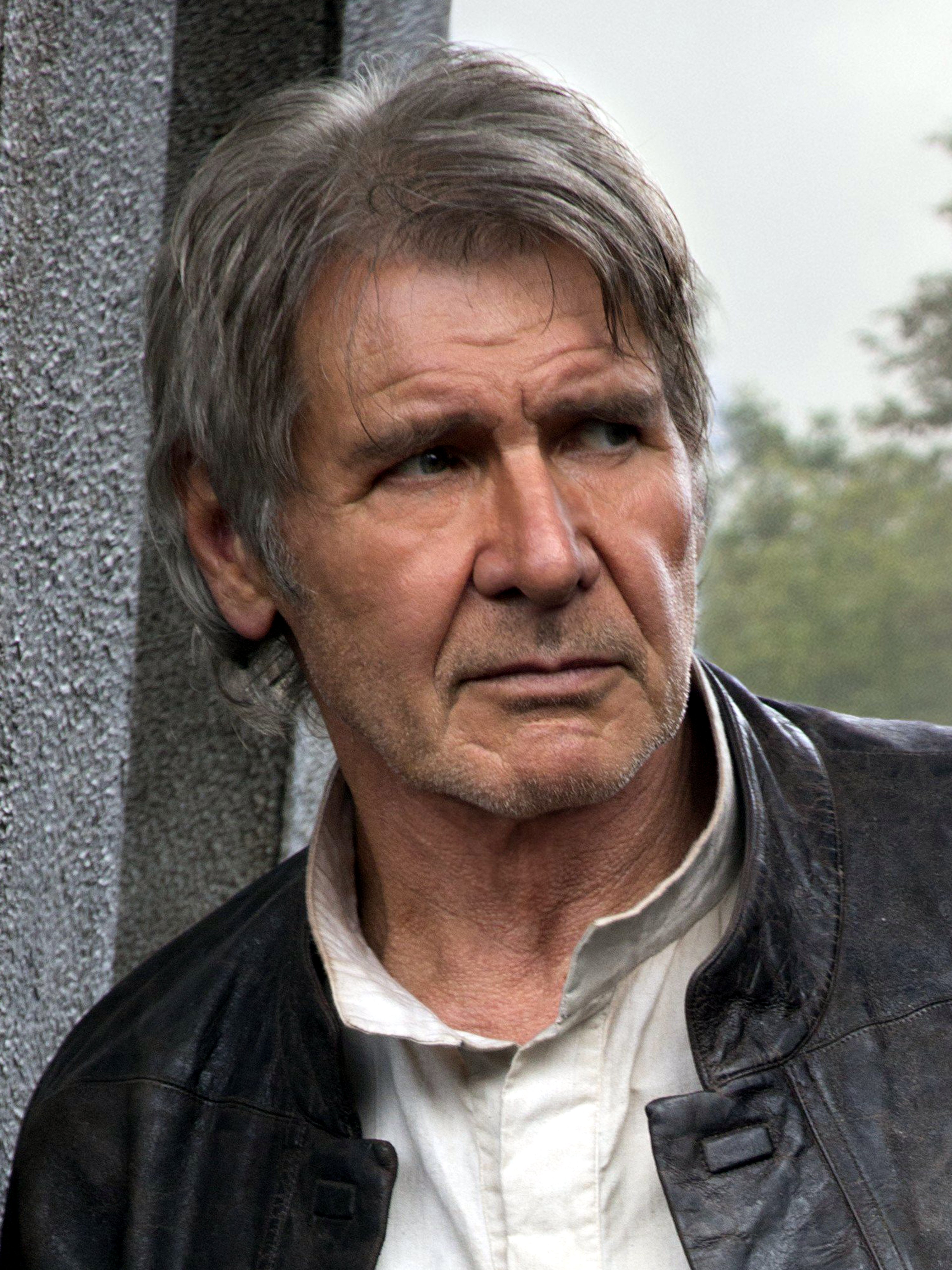 An older man with gray hair and a weathered face, wearing a dark jacket and leaning against a wall or building.