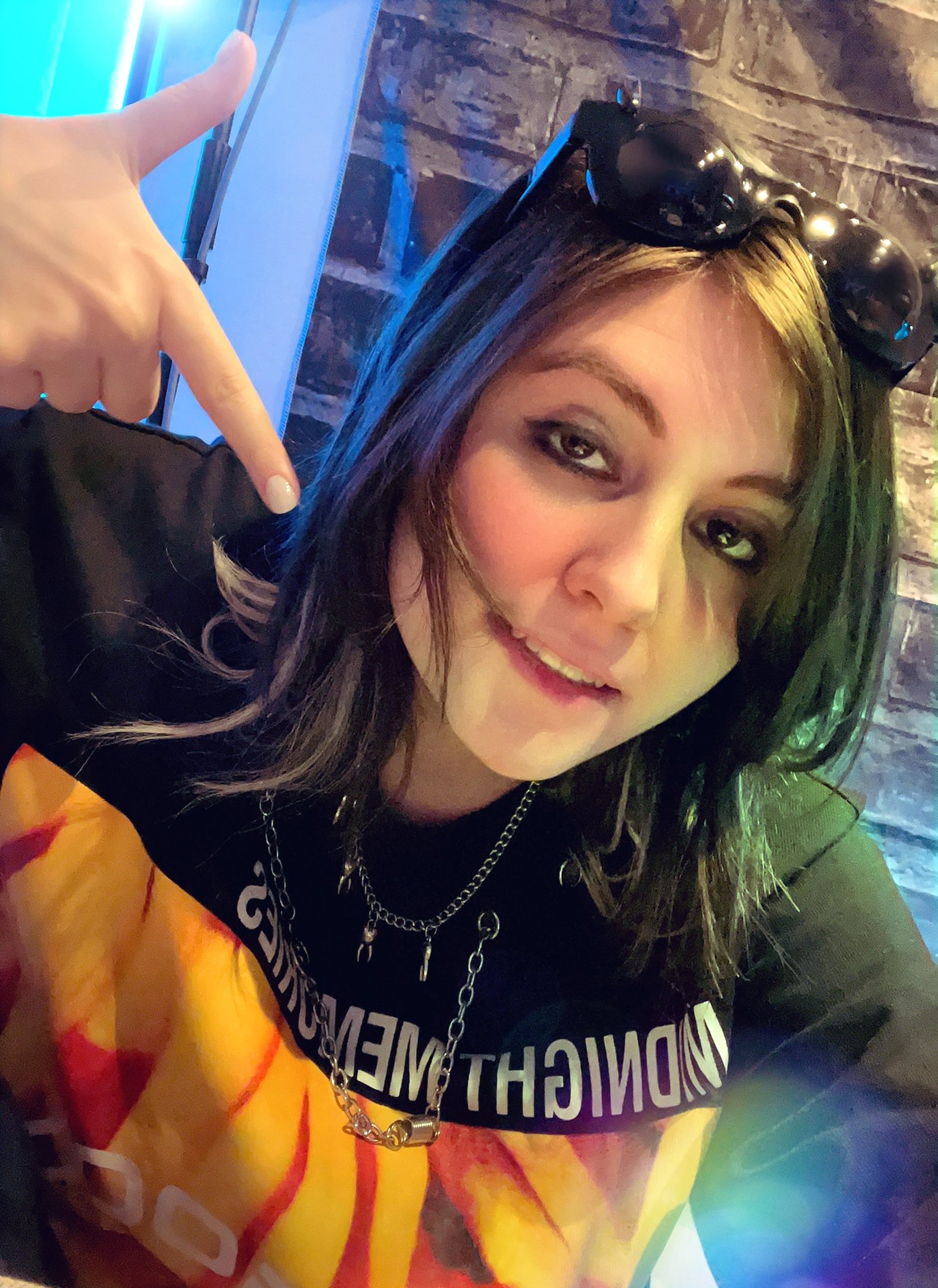 A young woman with dark hair and green highlights wearing a black shirt with a demon-like character design, looking at the camera with a confident expression.