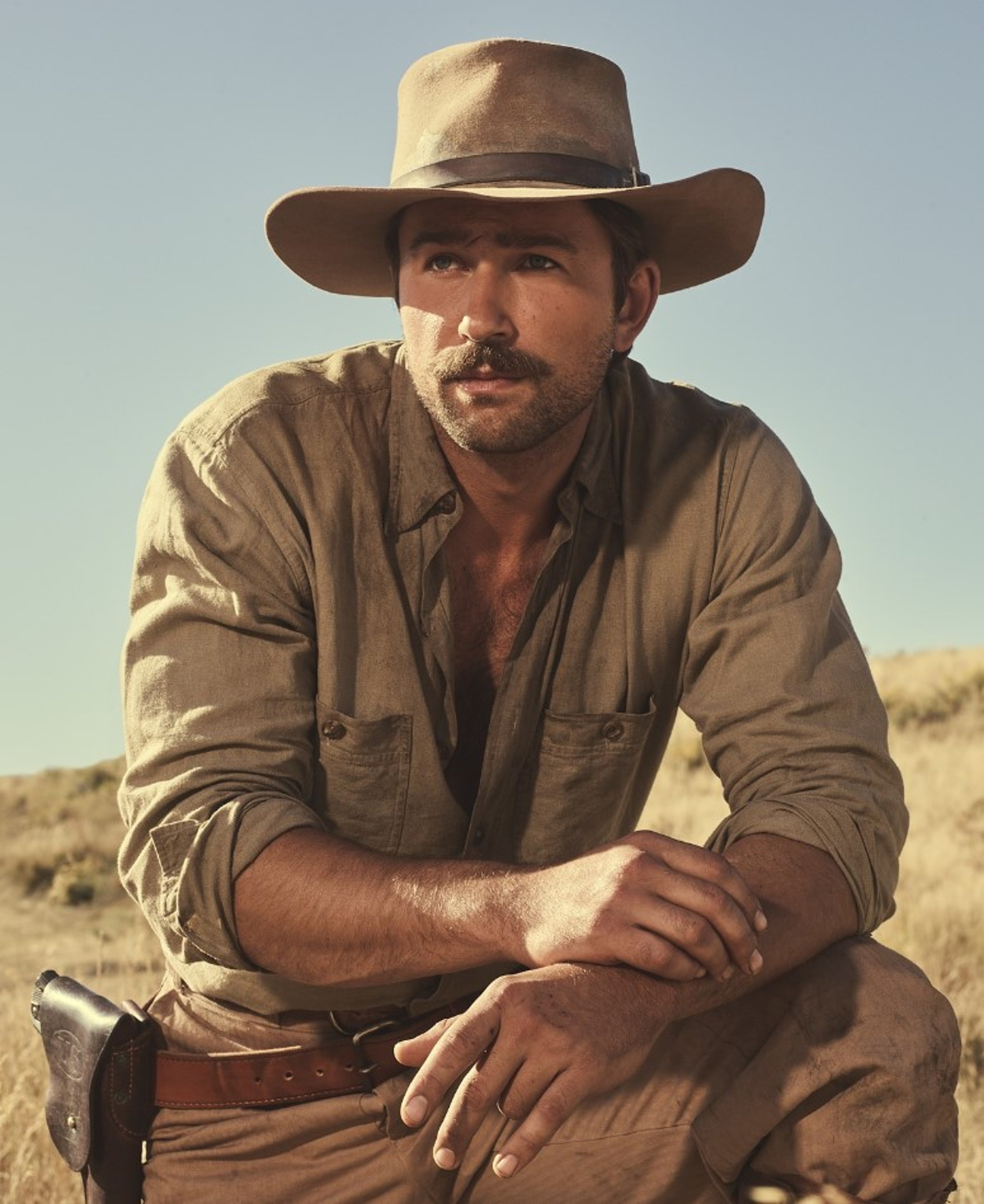 A man with a tan hat and shirt, a thick mustache and beard, and piercing blue eyes