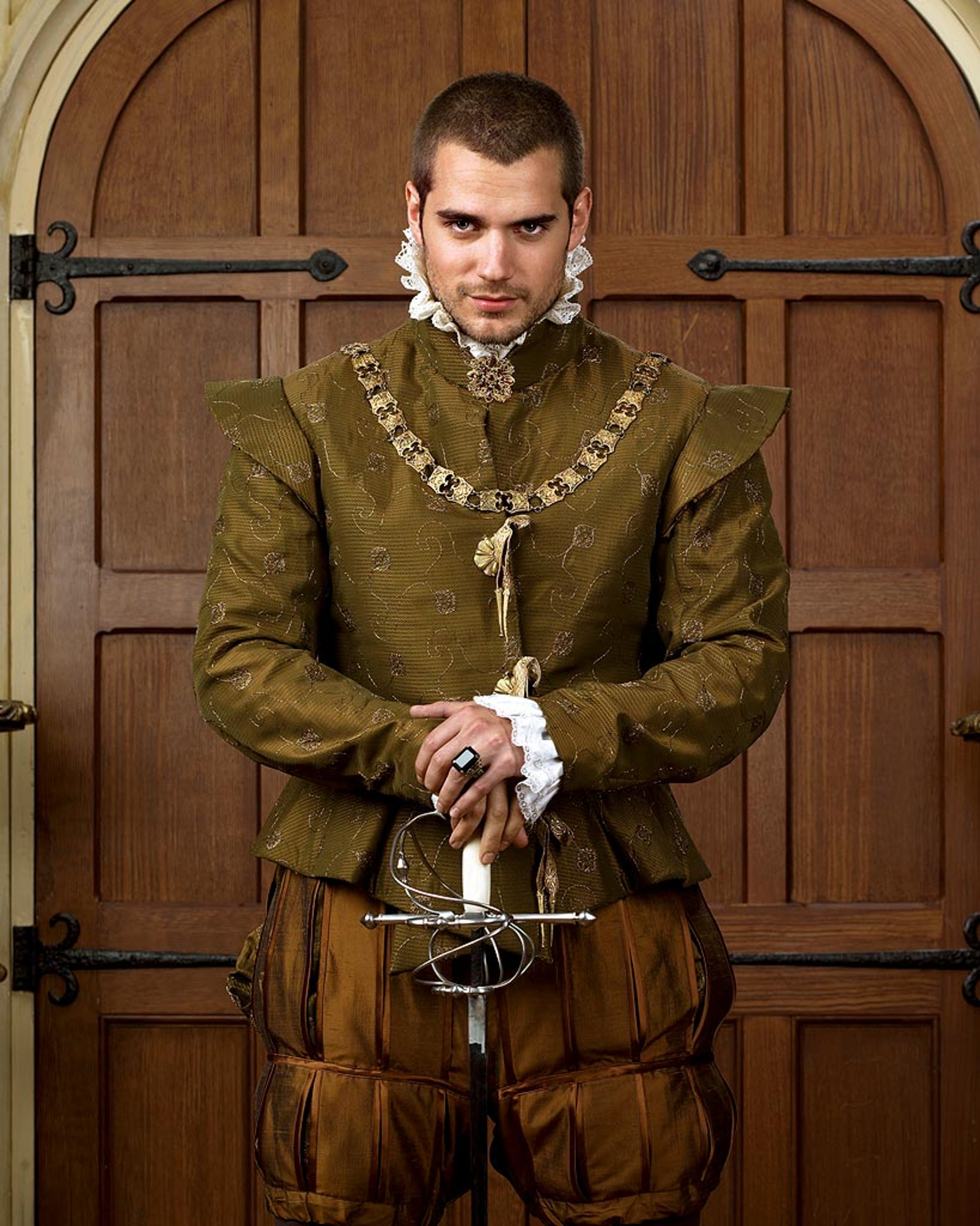 A man in a green military jacket holding a sword in front of a wooden door