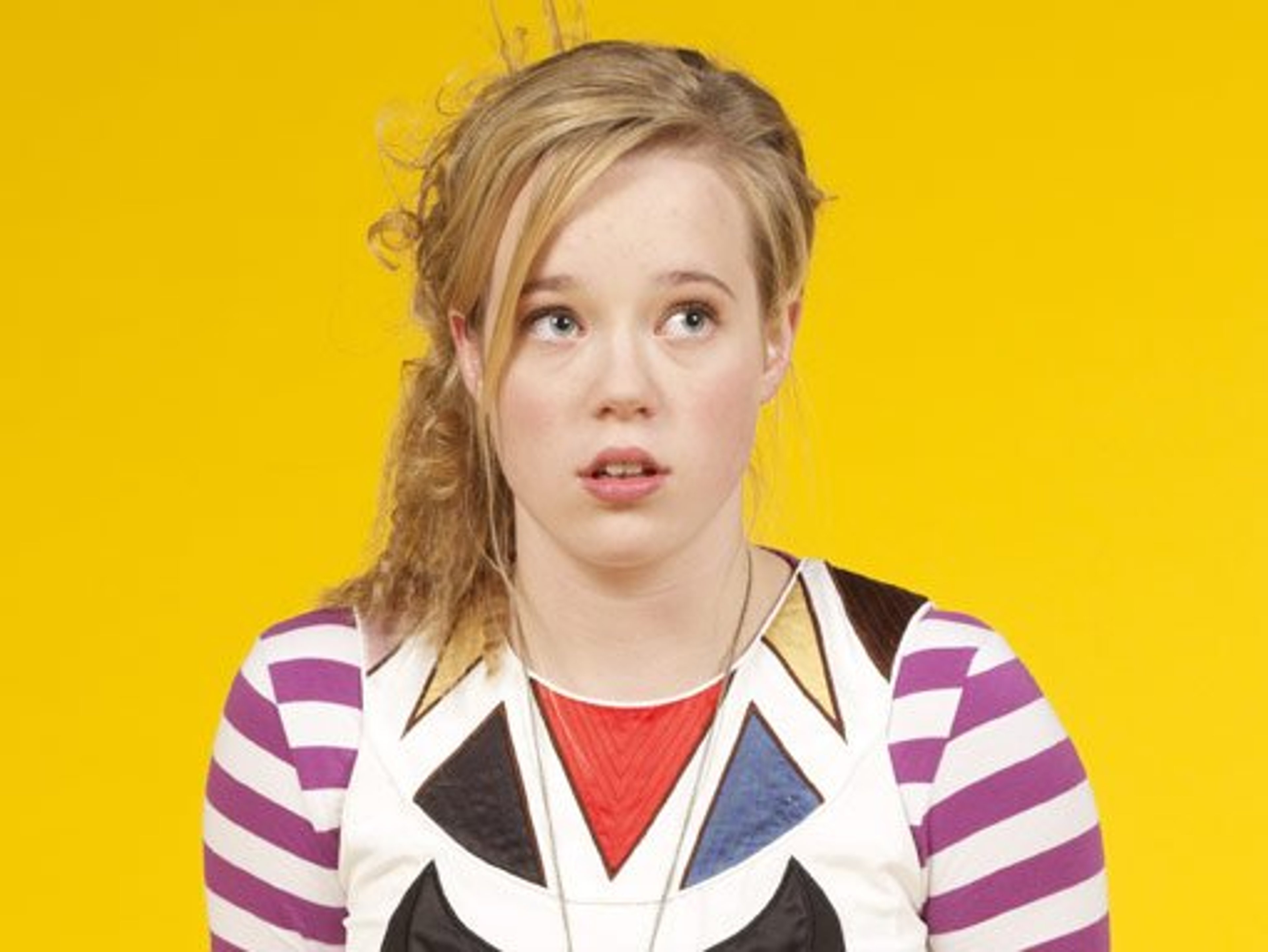 A young woman with blonde, curly hair wearing a striped shirt against a yellow background