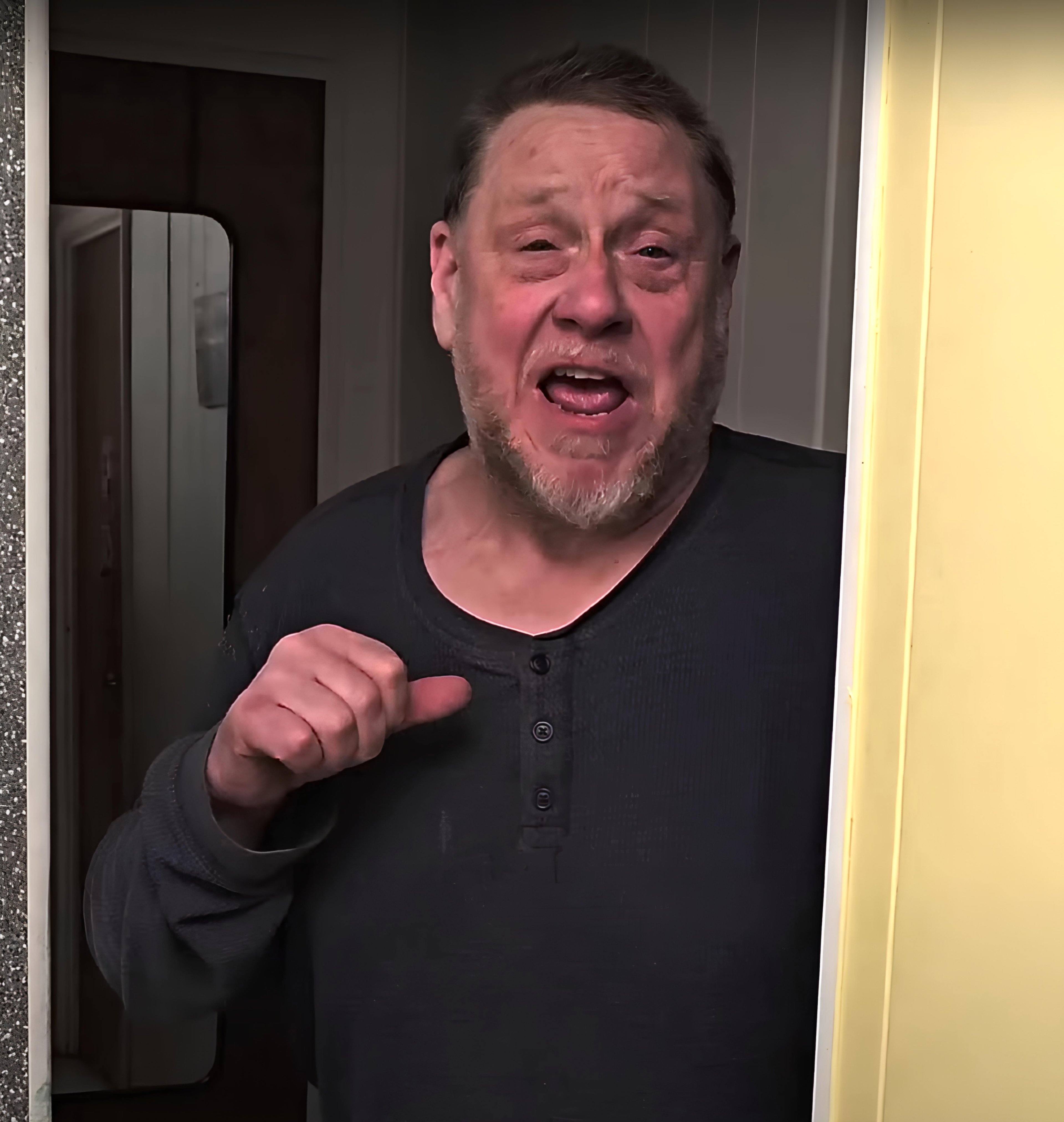 A middle-aged man with a beard and glasses, wearing a black shirt, with an angry, scowling expression on his face.