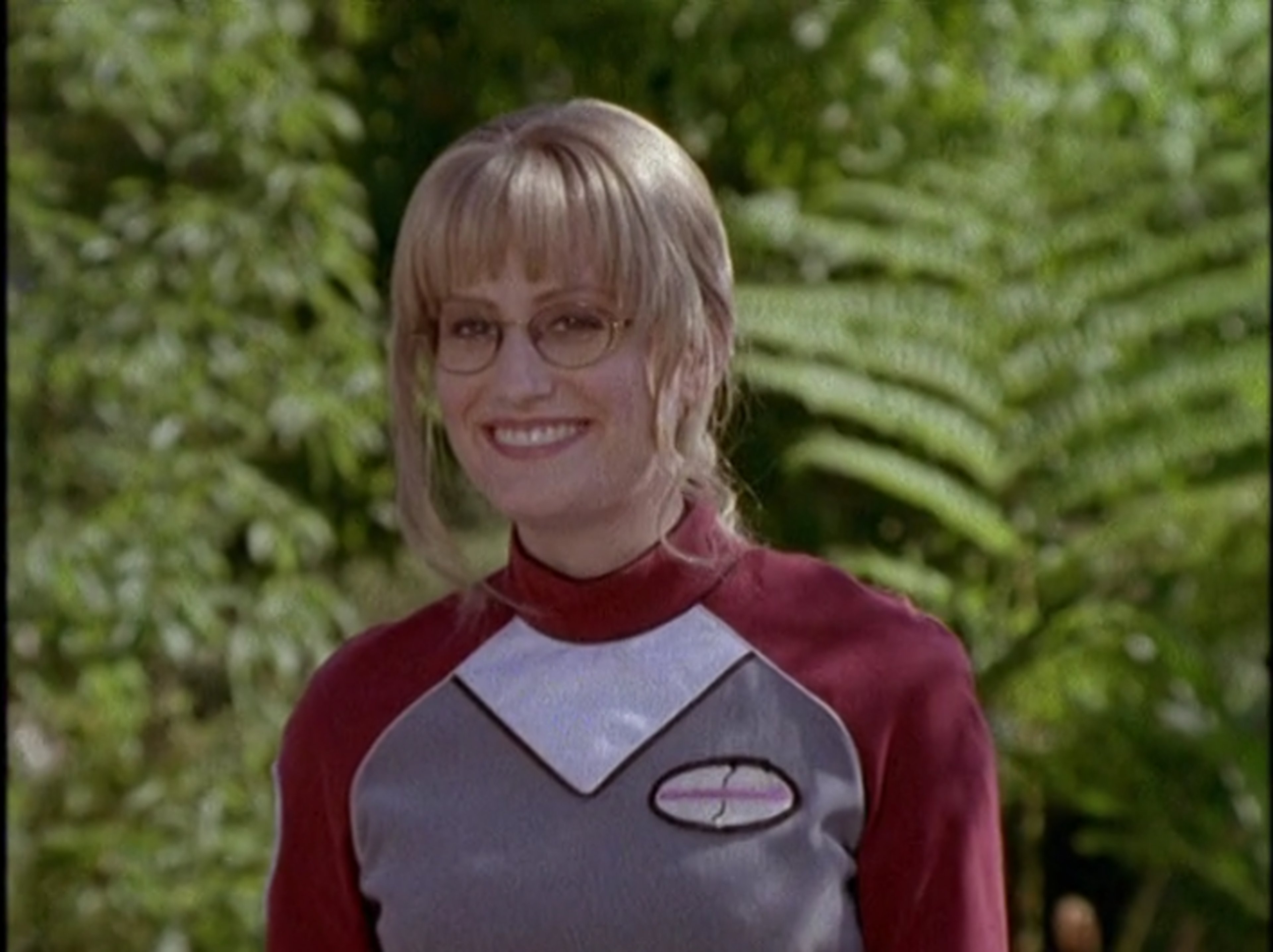 A woman in a red and gray uniform with blonde hair and glasses, standing in a natural outdoor setting.
