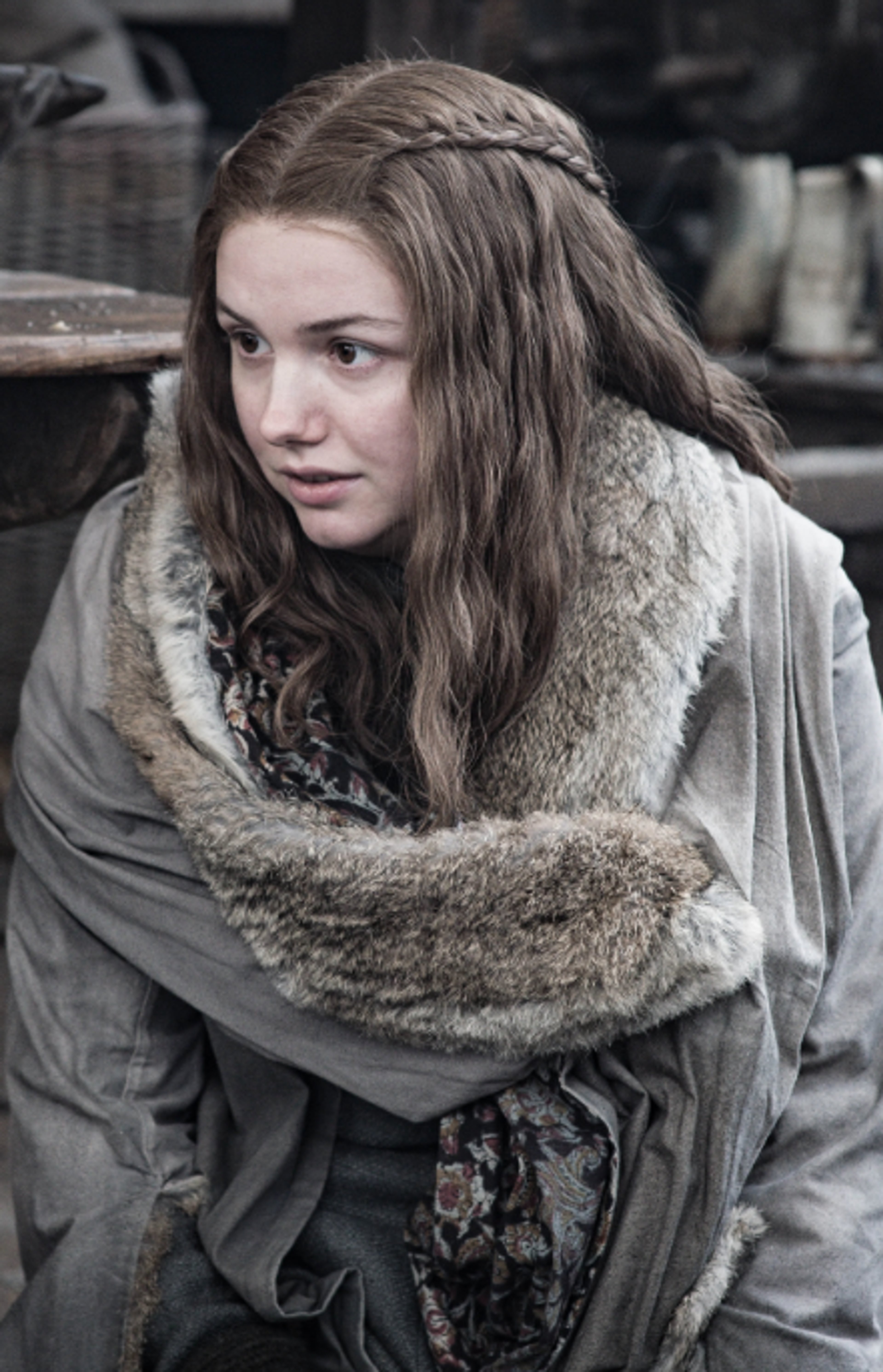 A young woman with long, wavy brown hair wearing a fur-lined cloak in a rustic setting