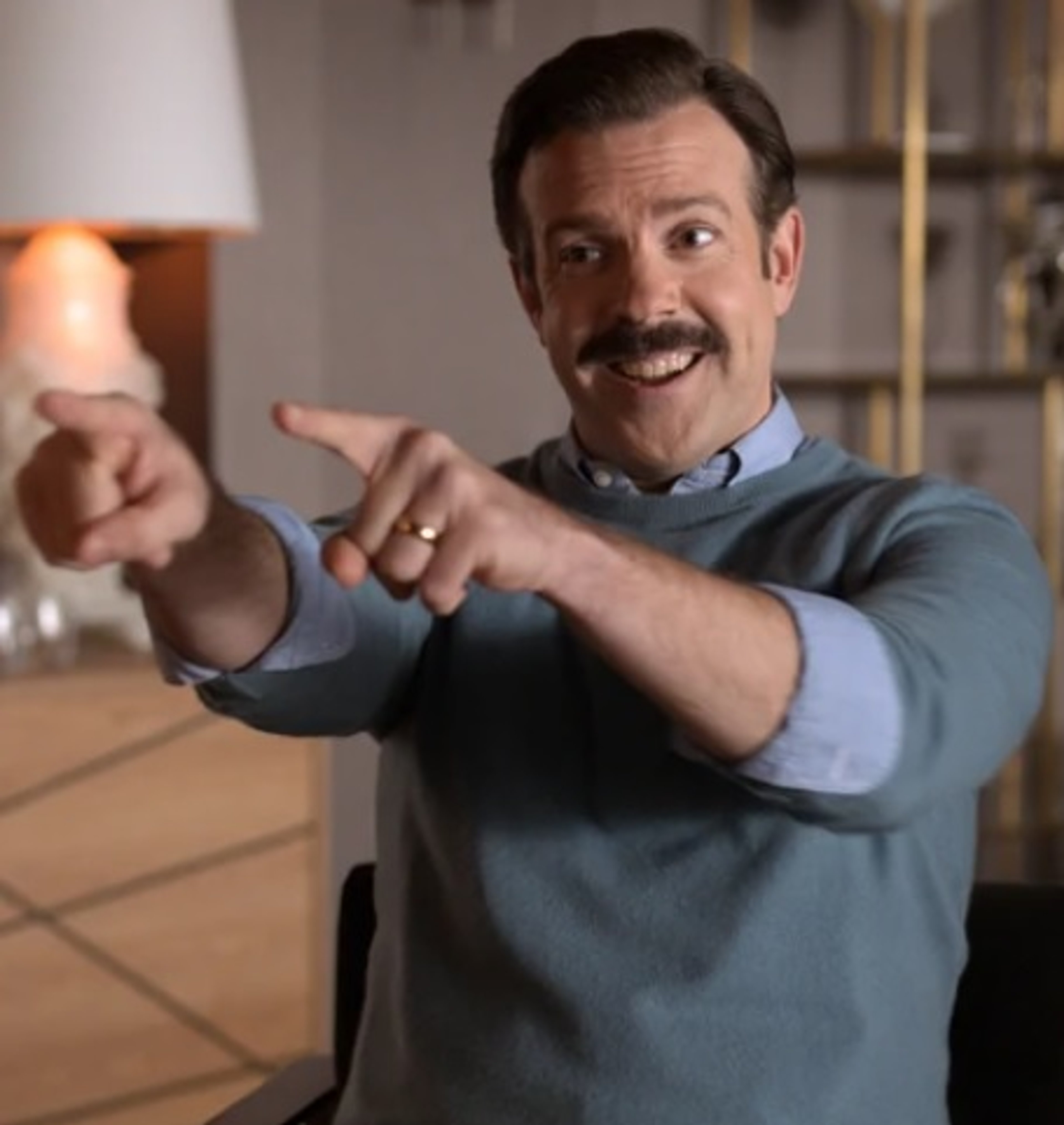 A middle-aged man with a mustache and beard, wearing a blue shirt, gesturing and speaking in a friendly manner.