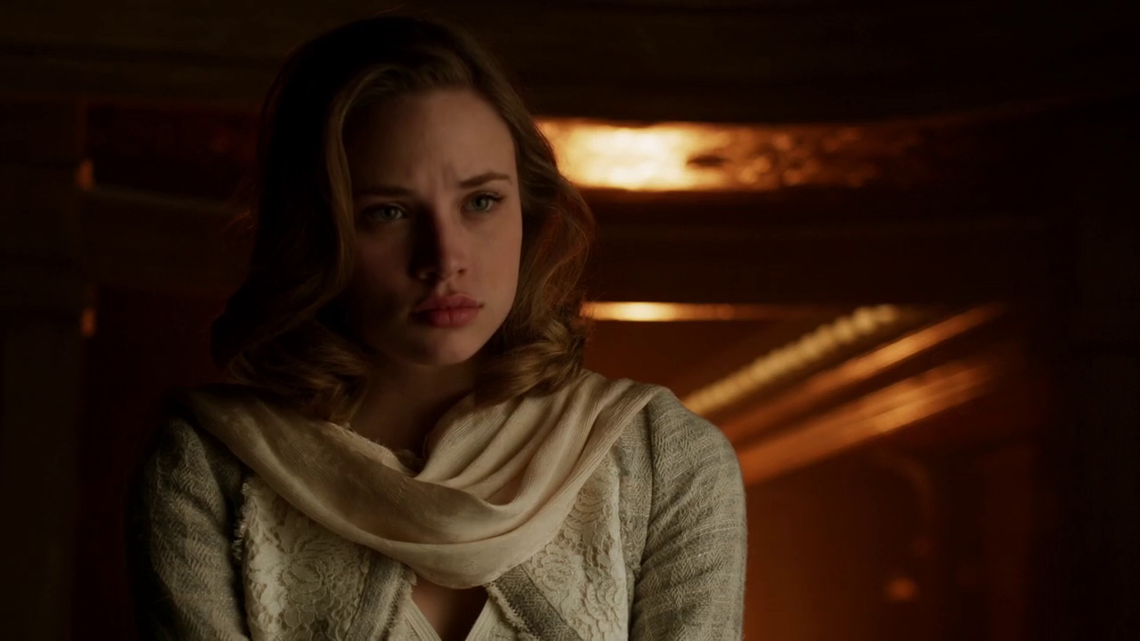 A young woman with dark hair wearing a light-colored scarf or shawl, with a serious expression on her face in a dimly lit indoor setting.