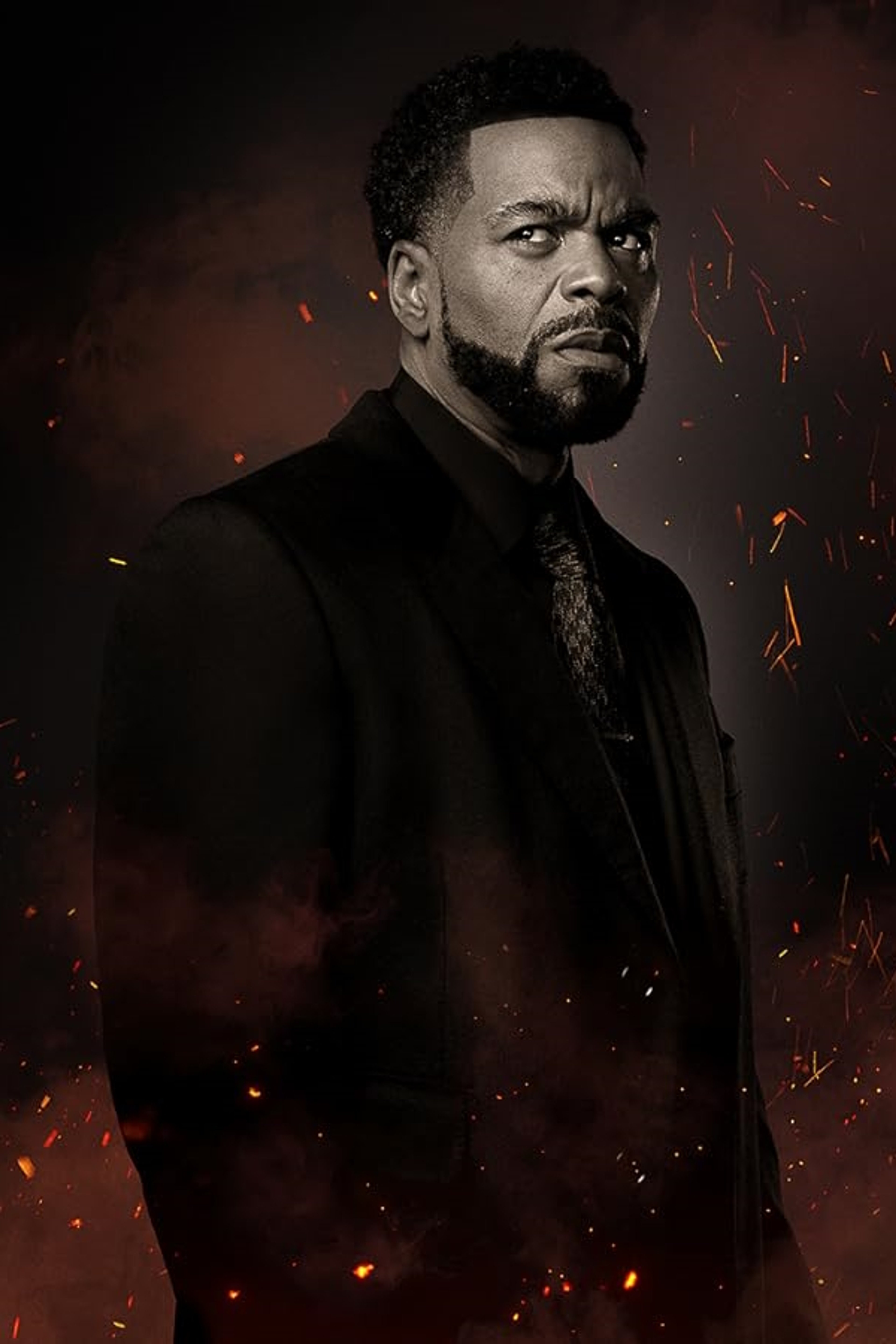 A serious-looking middle-aged Black man in a dark suit and tie against a dark, fiery background