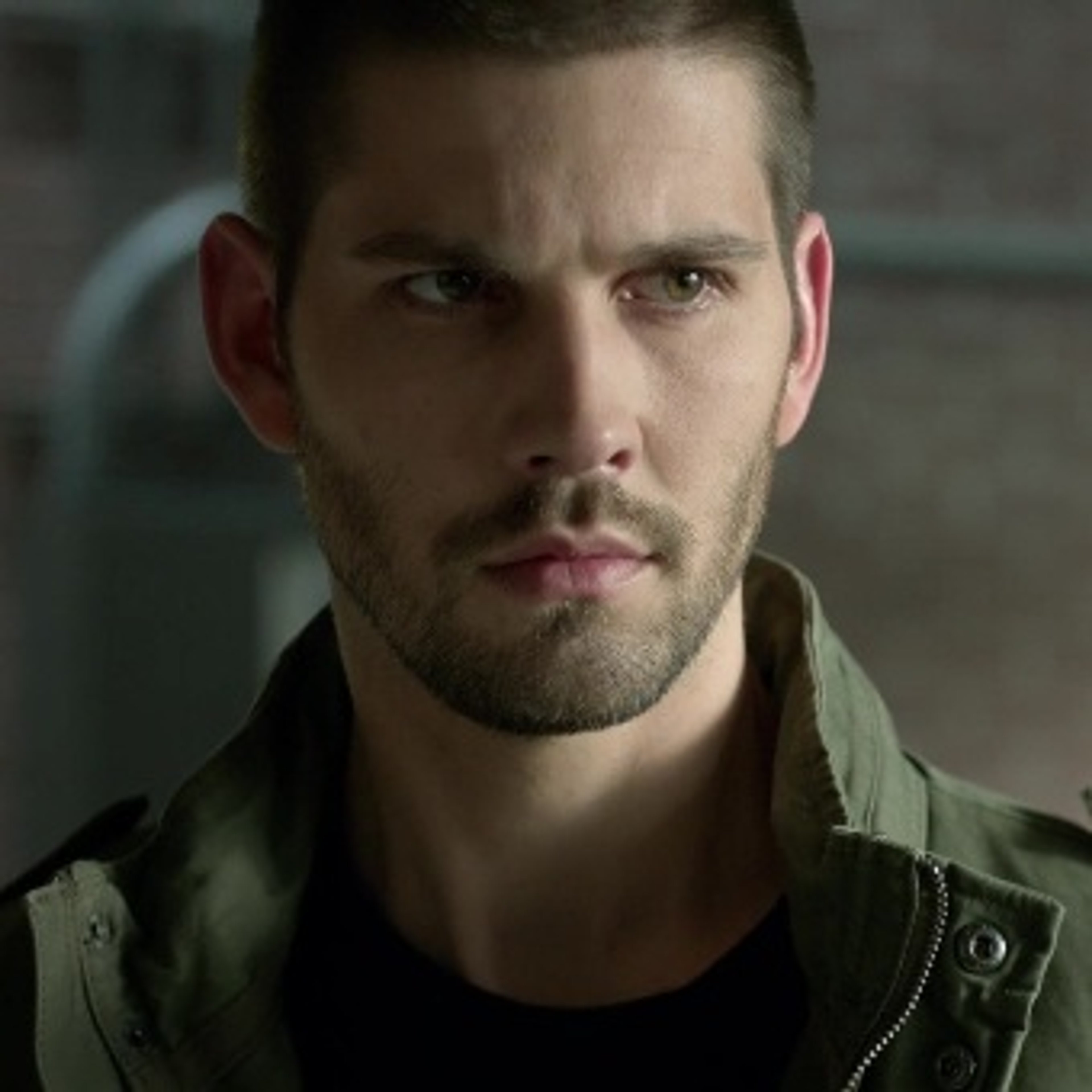 A man with dark brown hair and intense brown eyes wearing a green military jacket