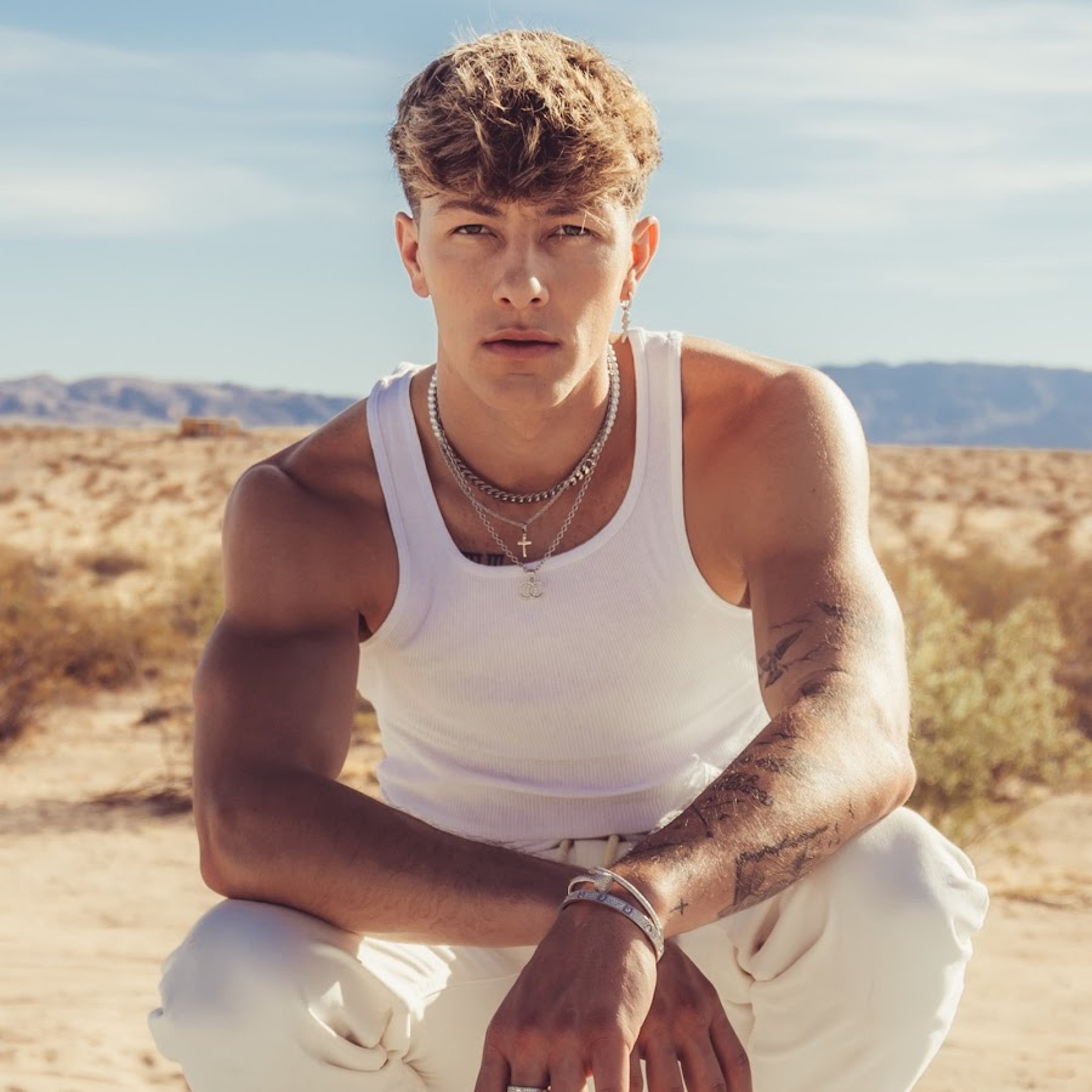 A young, muscular man with curly blonde hair posing outdoors
