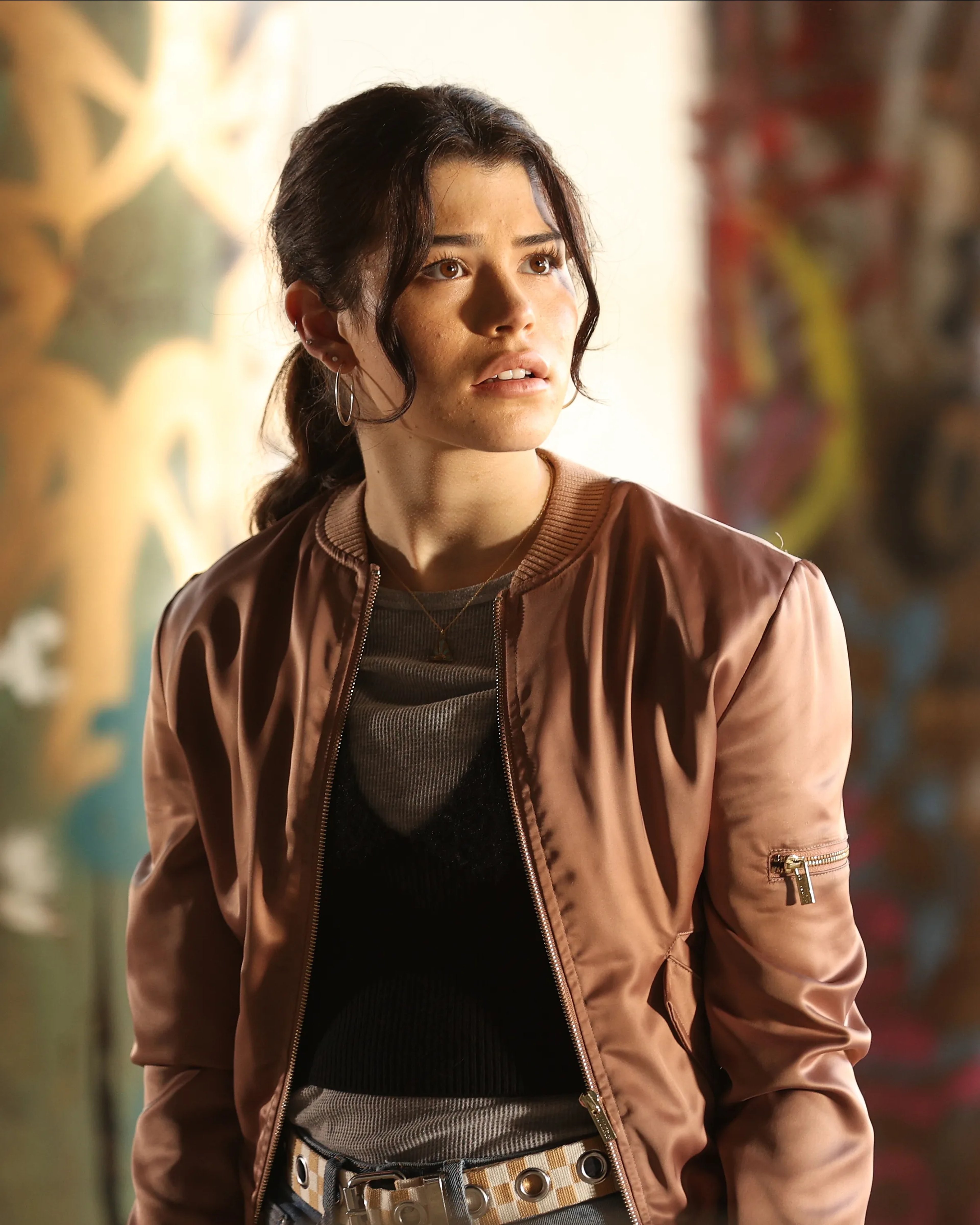 A young woman with dark hair wearing a pink jacket in an urban setting