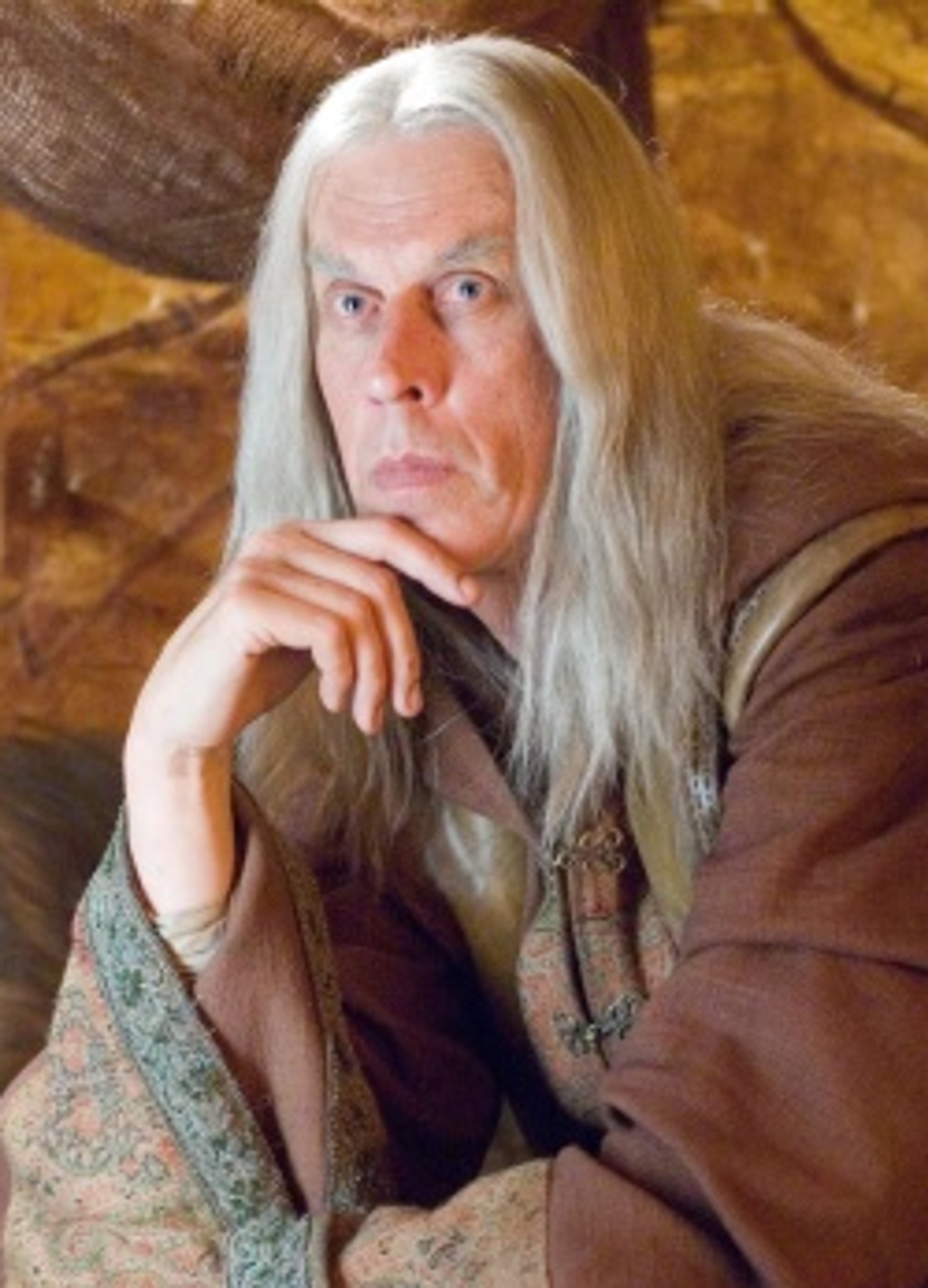 An elderly man with long white hair and a beard, wearing tan robes and looking thoughtful.