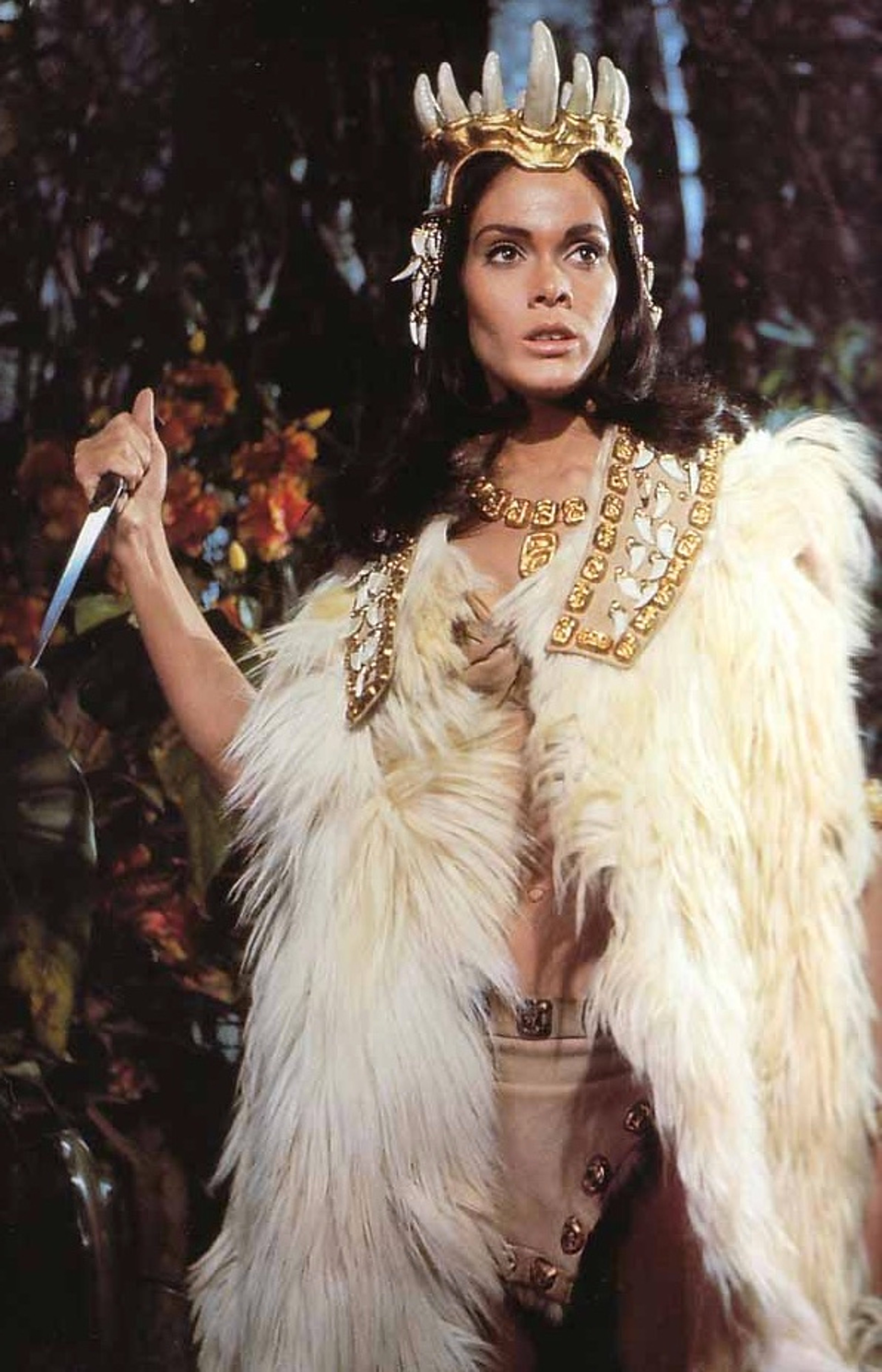 A woman with dark hair wearing an elaborate headdress and jewelry, with a fur-trimmed cape or cloak, standing with a commanding presence.