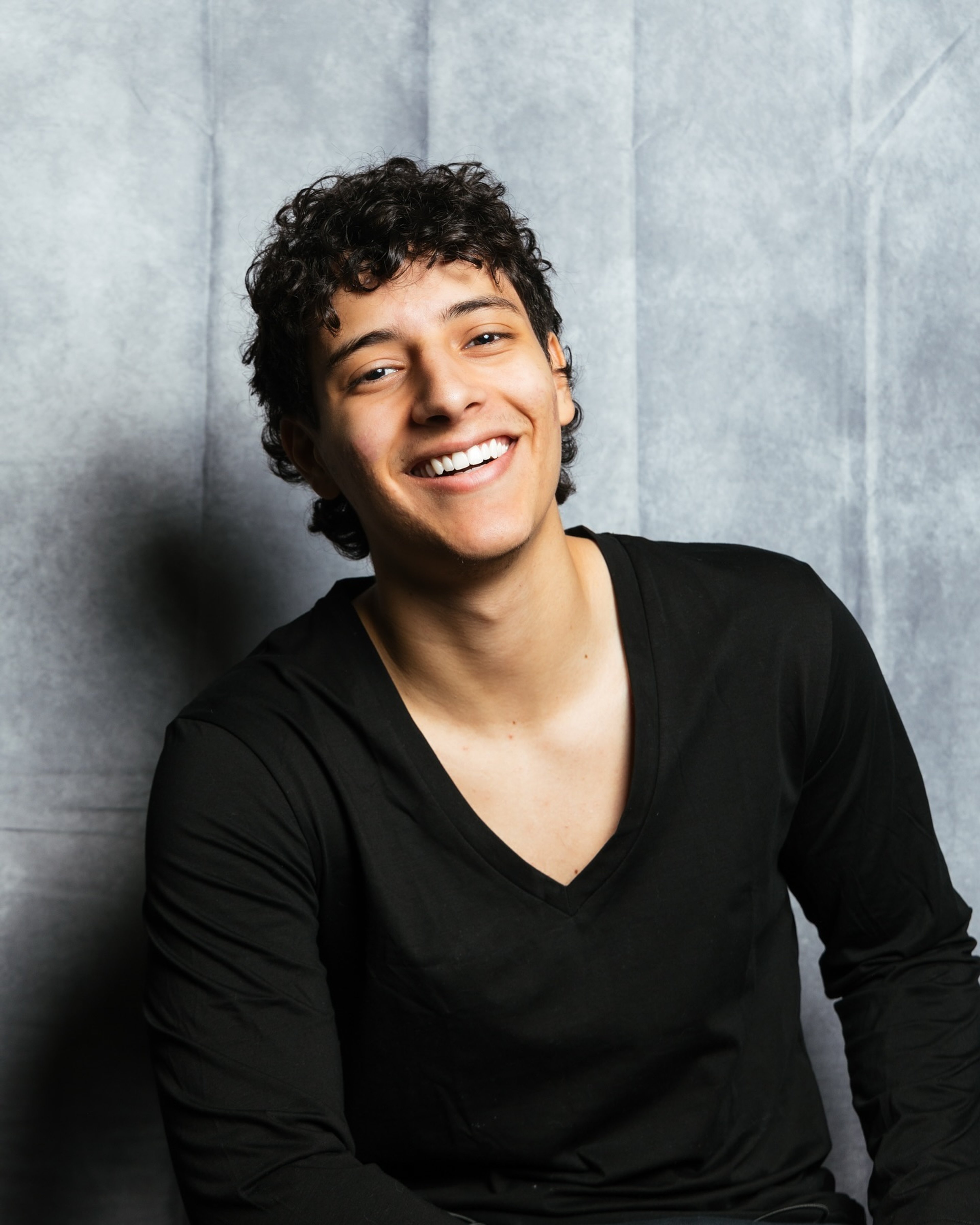 A young man with curly dark hair and a friendly smile, wearing a black v-neck shirt.