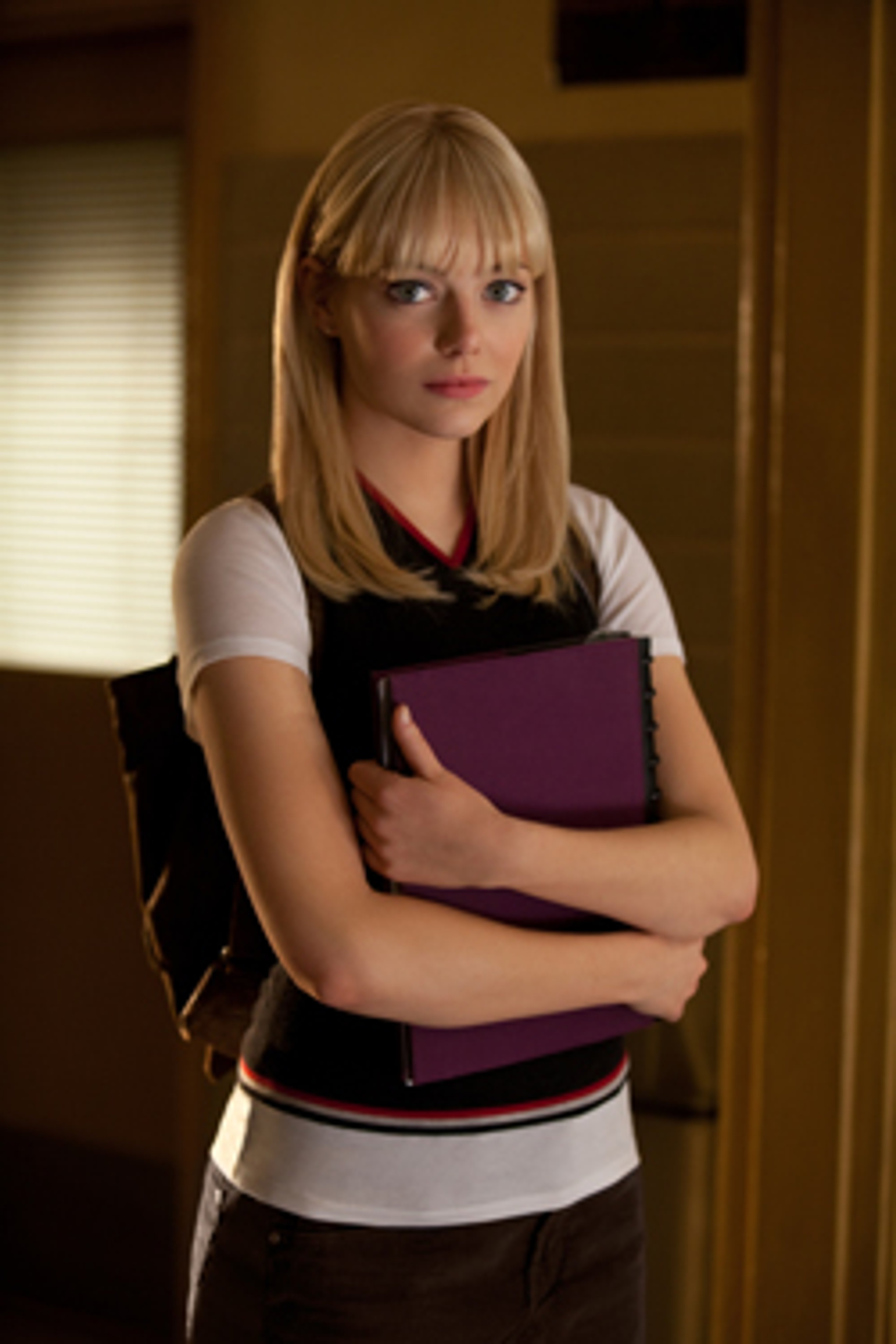 A young woman with blonde hair and blue eyes, wearing a white shirt and black vest, holding a purple folder or notebook.