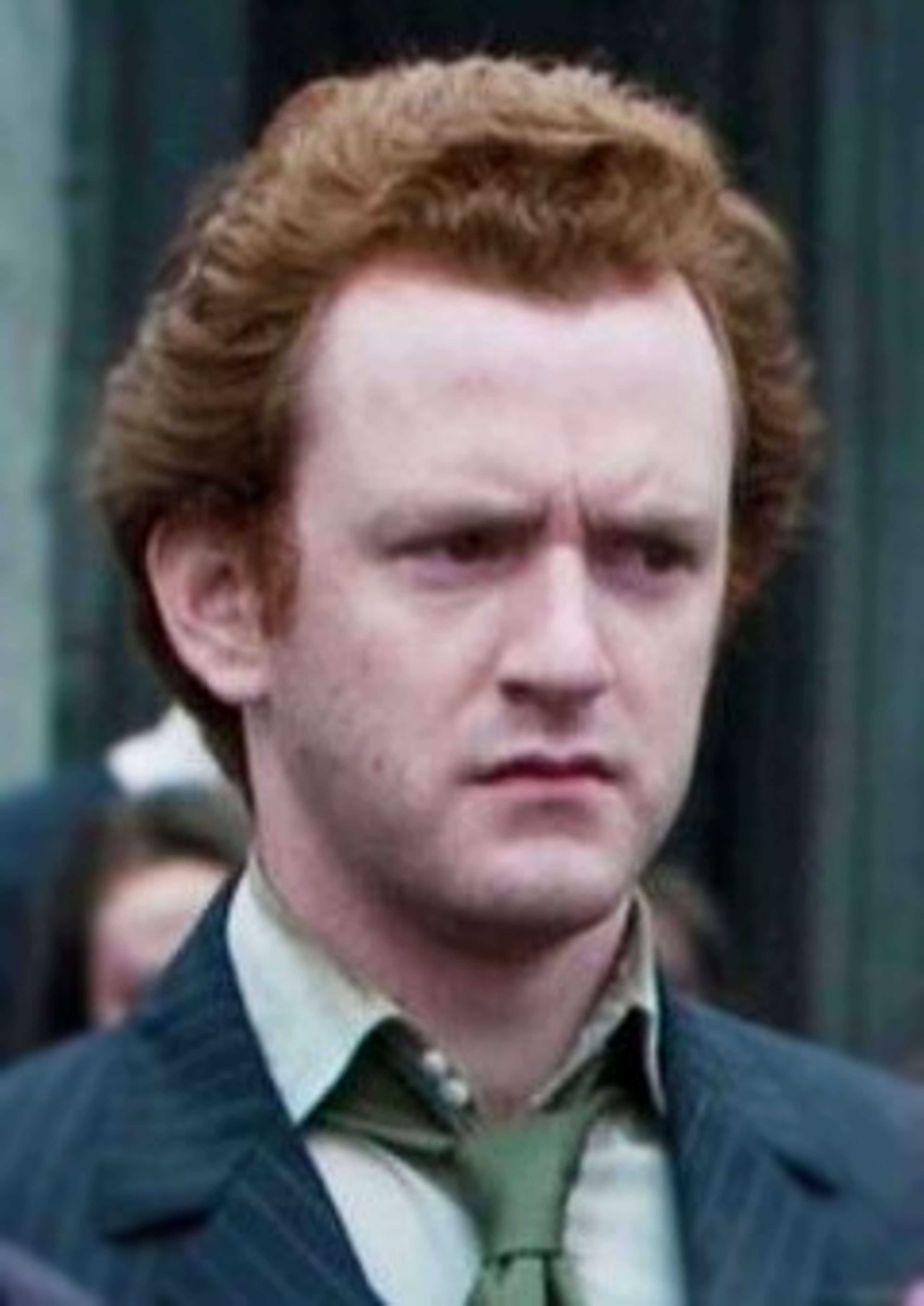 A middle-aged man with curly red hair wearing a green shirt and dark jacket