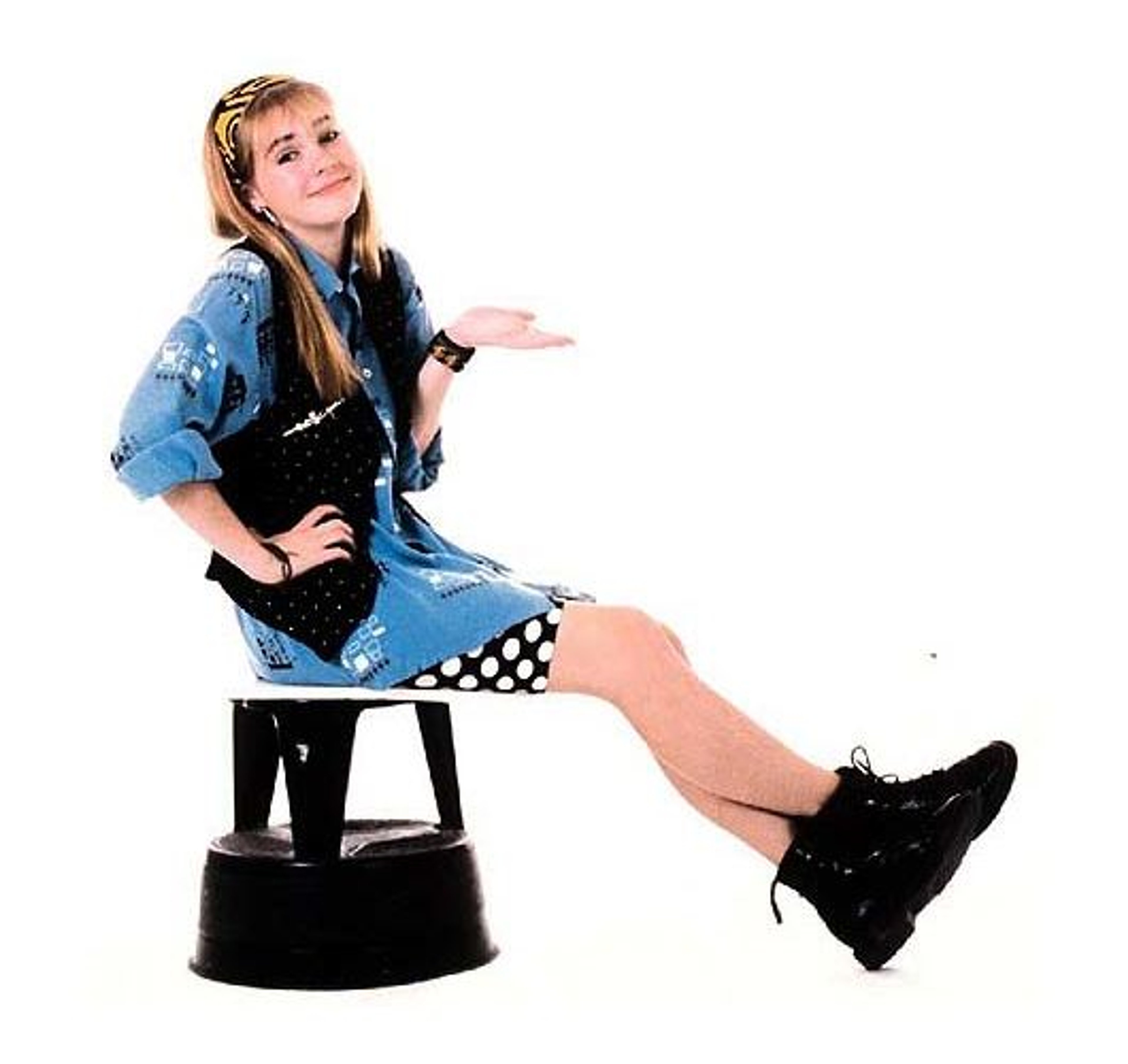A teenage girl with long blonde hair sitting on a stool, wearing a denim jacket, checkered skirt, and black boots.