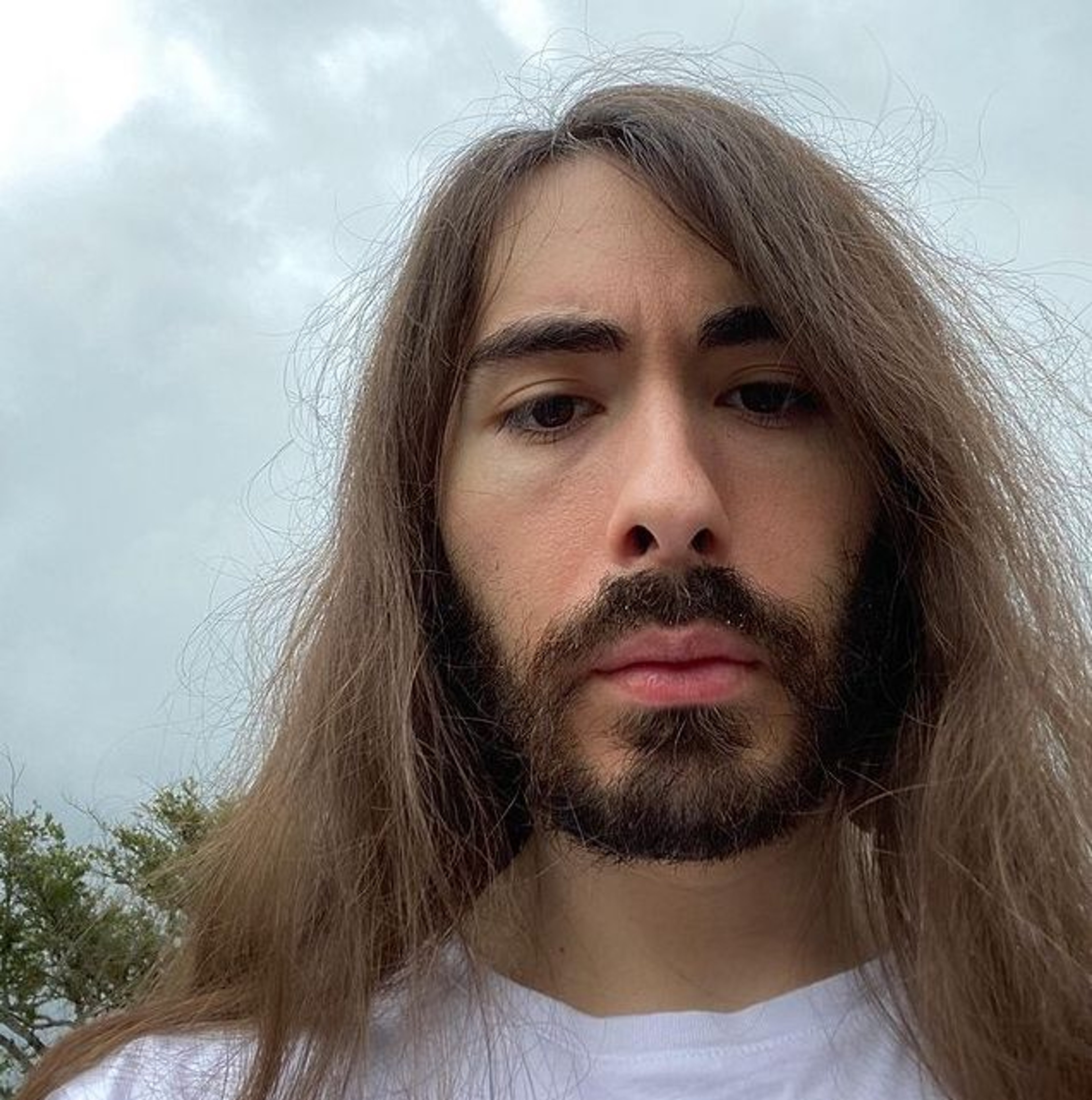 A man with long dark hair and a beard wearing a white t-shirt, looking serious