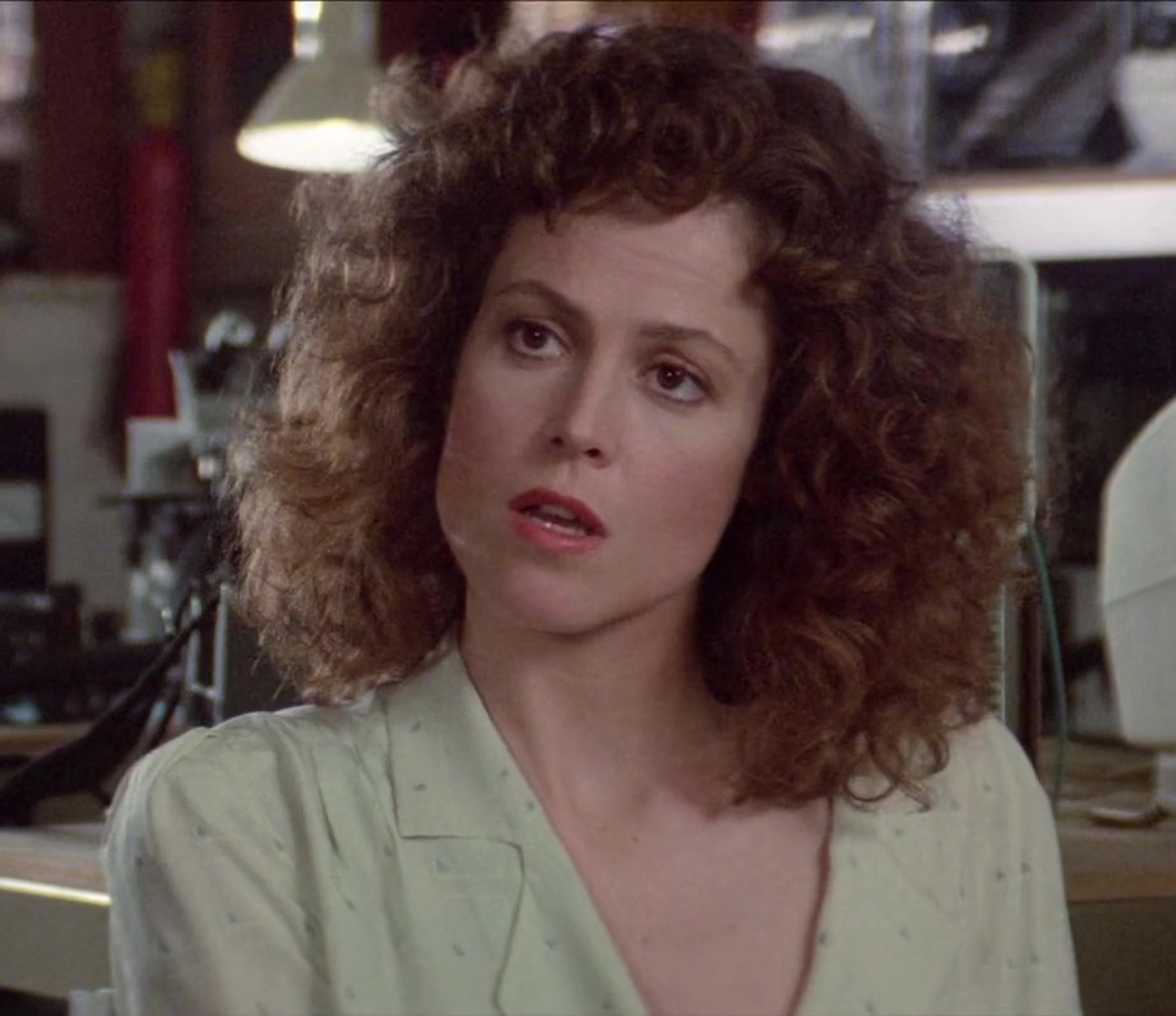 A woman with curly brown hair wearing a light-colored jacket in an indoor setting