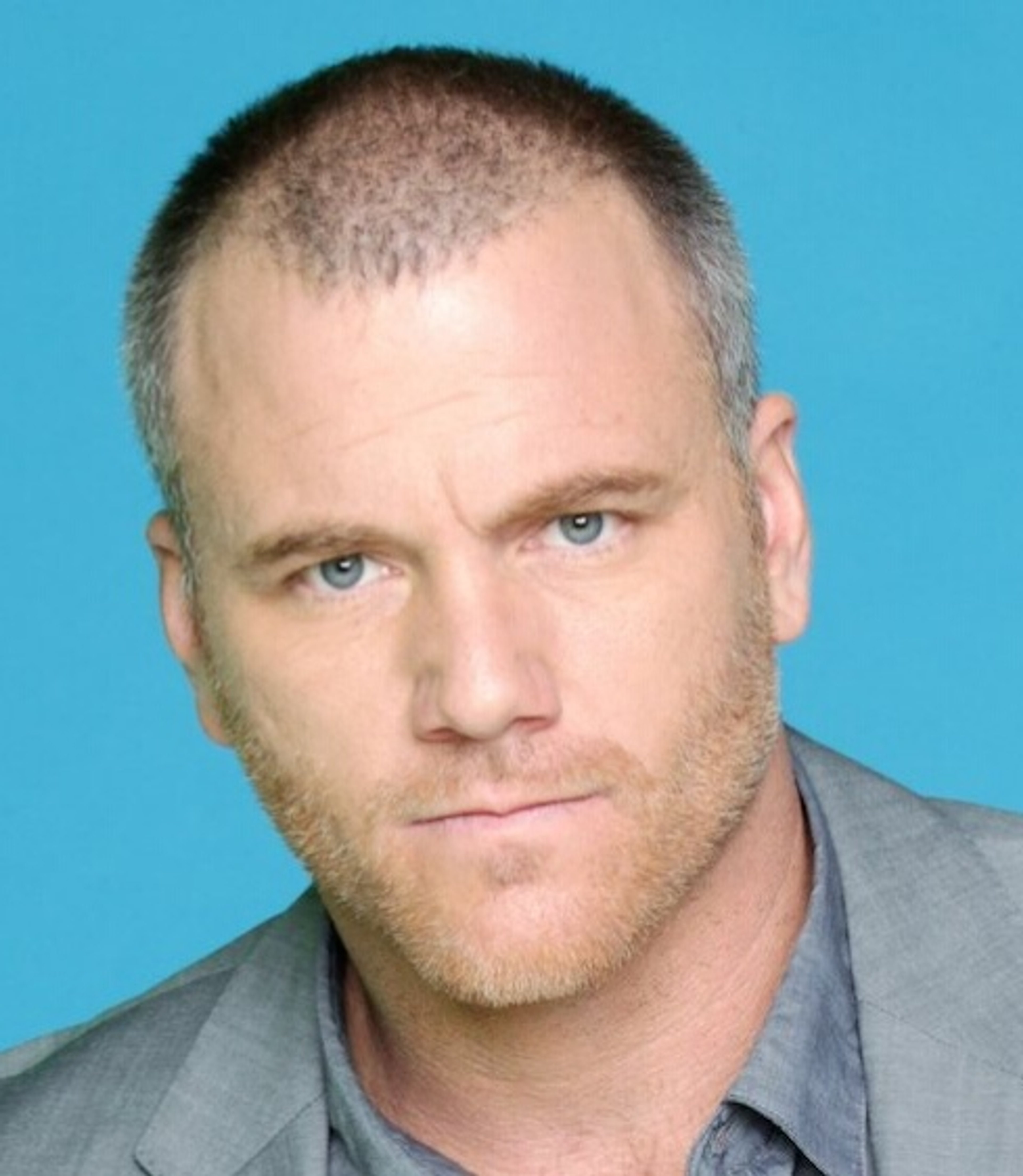 A middle-aged man with short brown hair and blue eyes, wearing a gray suit jacket