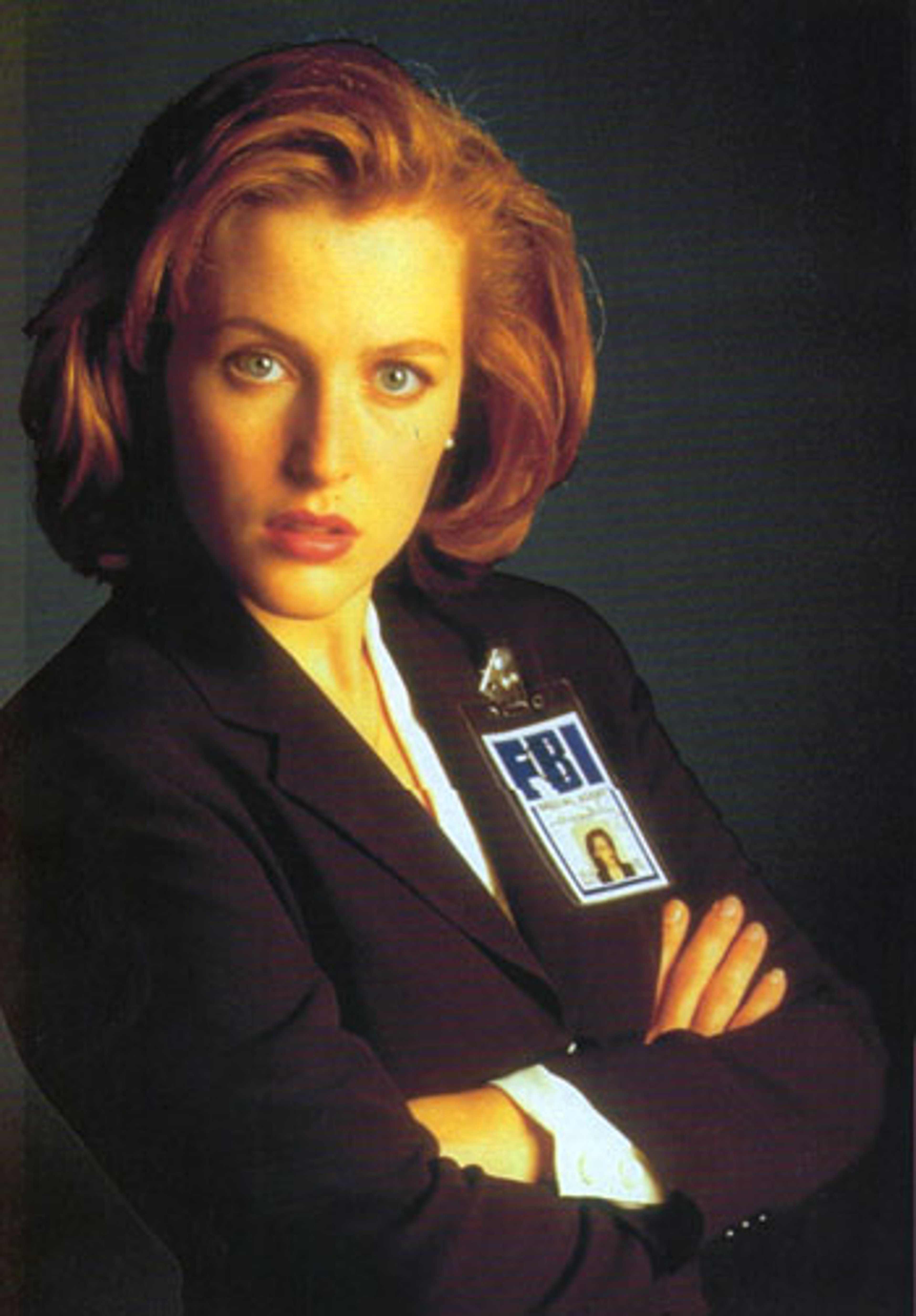 A woman with red hair wearing a dark suit and badge, standing with her arms crossed in a serious pose.