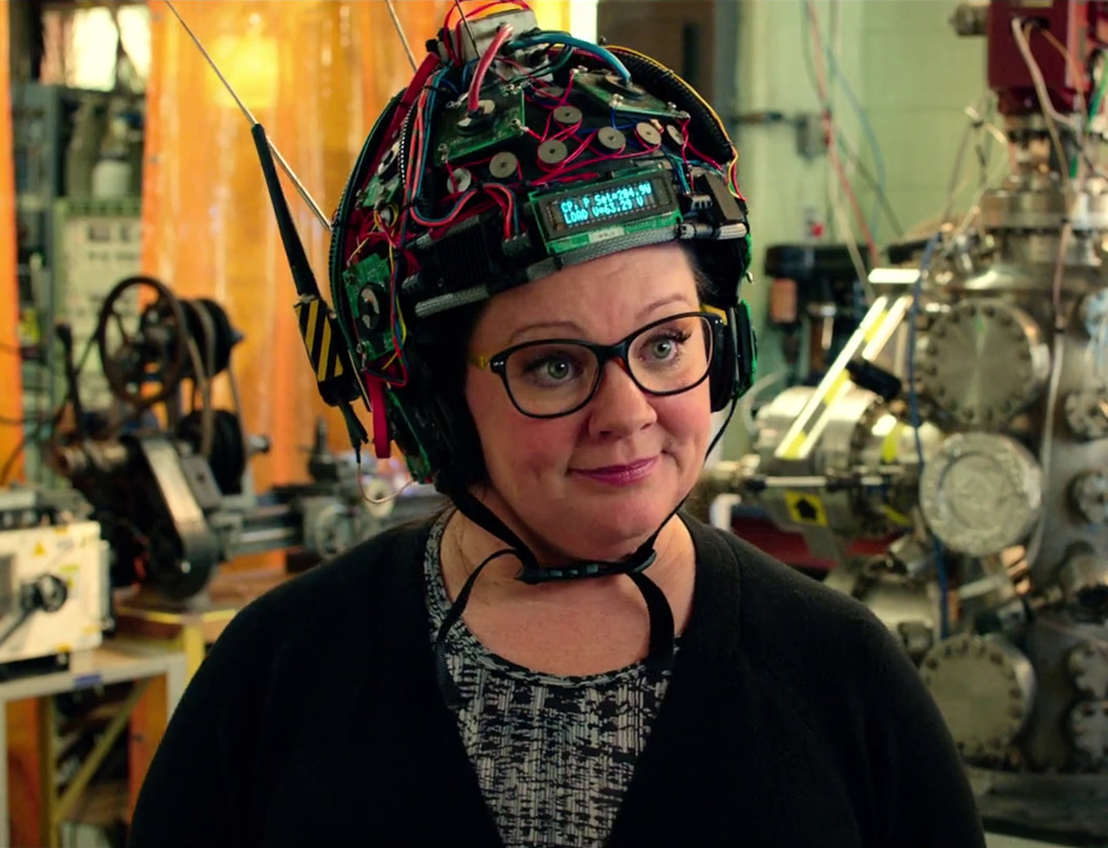 A middle-aged woman with short brown hair wearing glasses and a colorful patterned hat, standing in a workshop or laboratory setting.