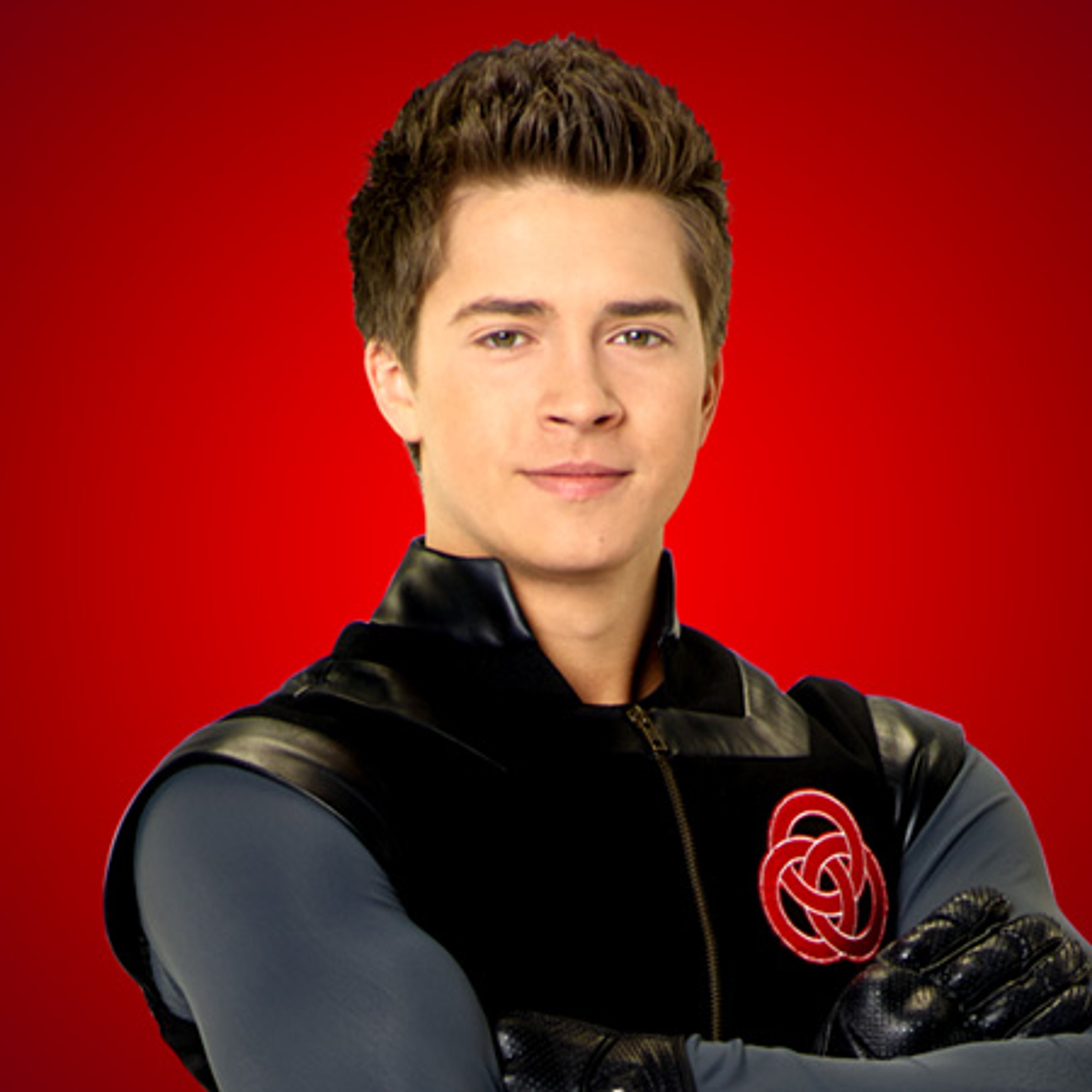 A young man with short brown hair wearing a black jacket with a red rose emblem on the chest, standing against a red background.