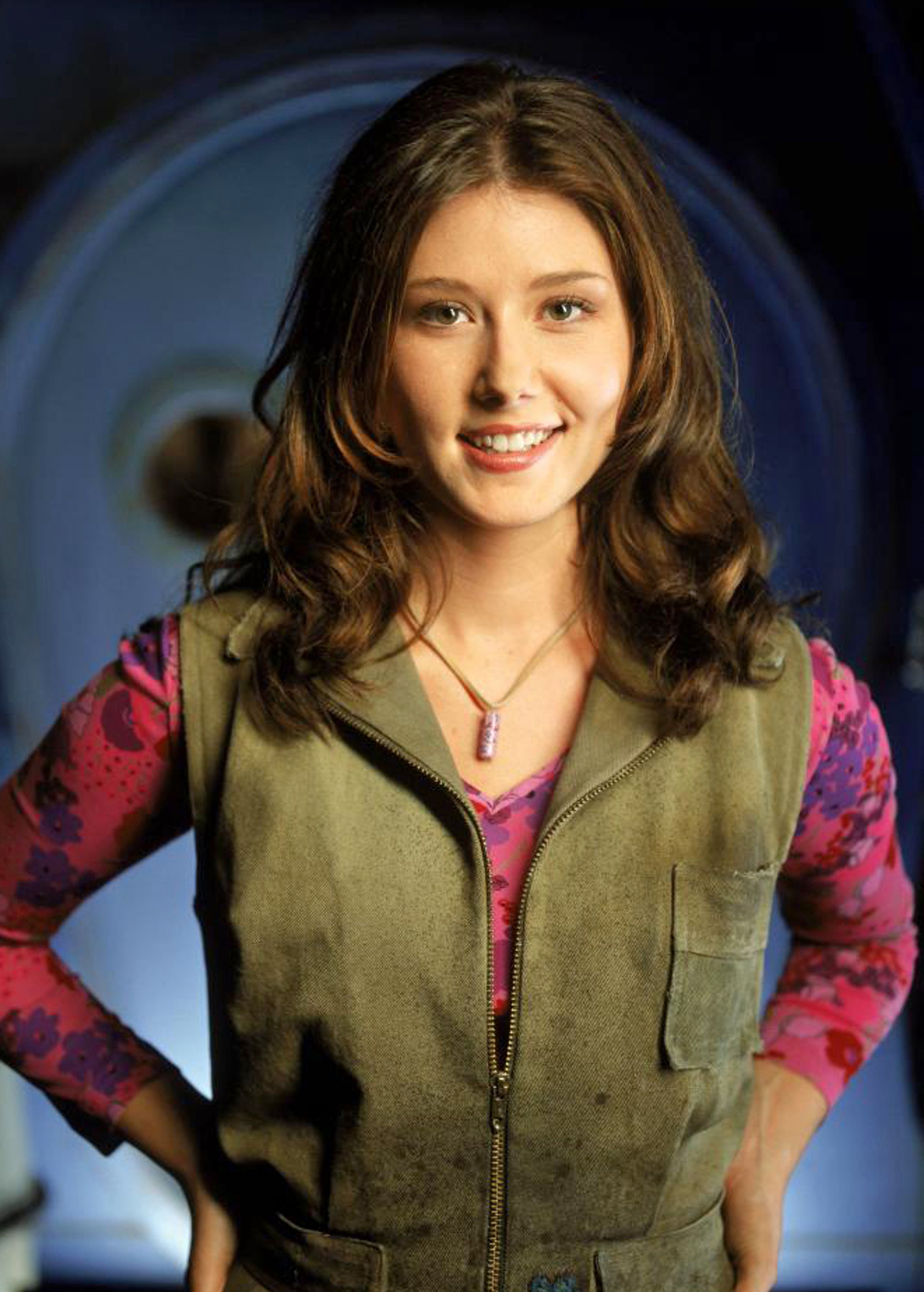 A young woman with long brown hair and a friendly smile, wearing a plaid shirt and green vest.