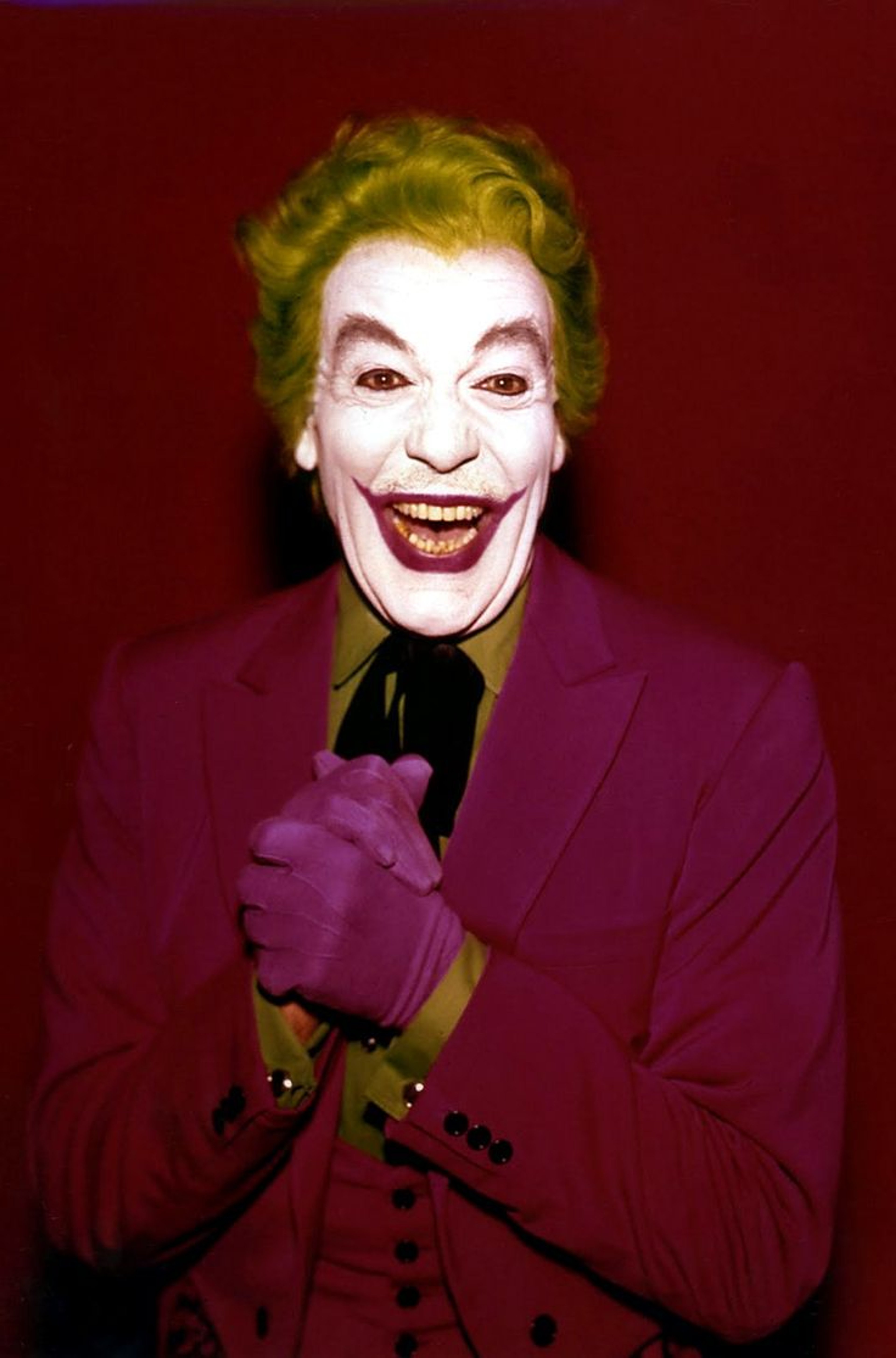 A man with a clownish appearance, wearing a purple suit and green wig, laughing maniacally.