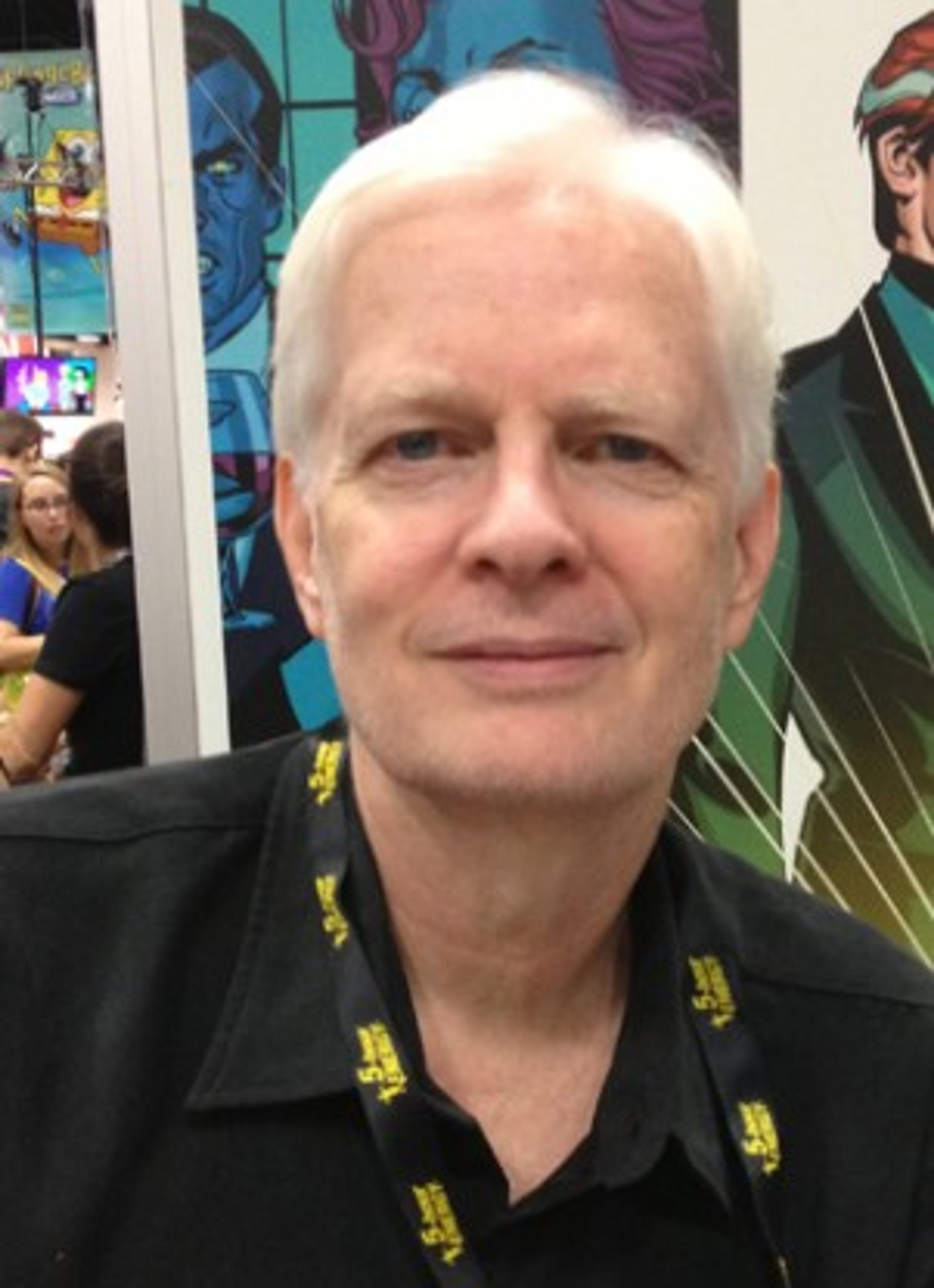 An older man with gray hair and a friendly expression, wearing a black shirt with a yellow pattern.