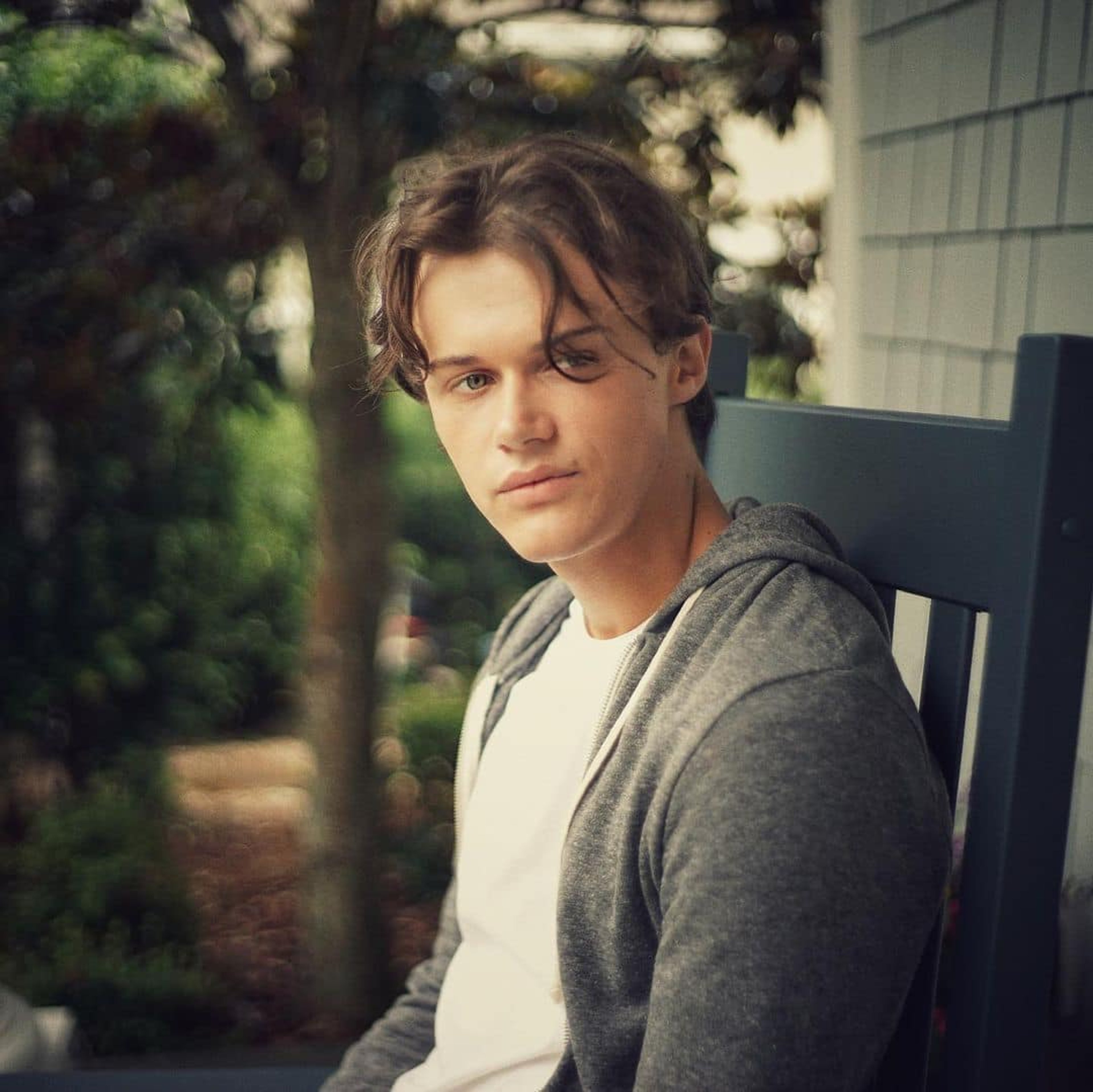 A young man with brown hair wearing a gray sweater, sitting outdoors.