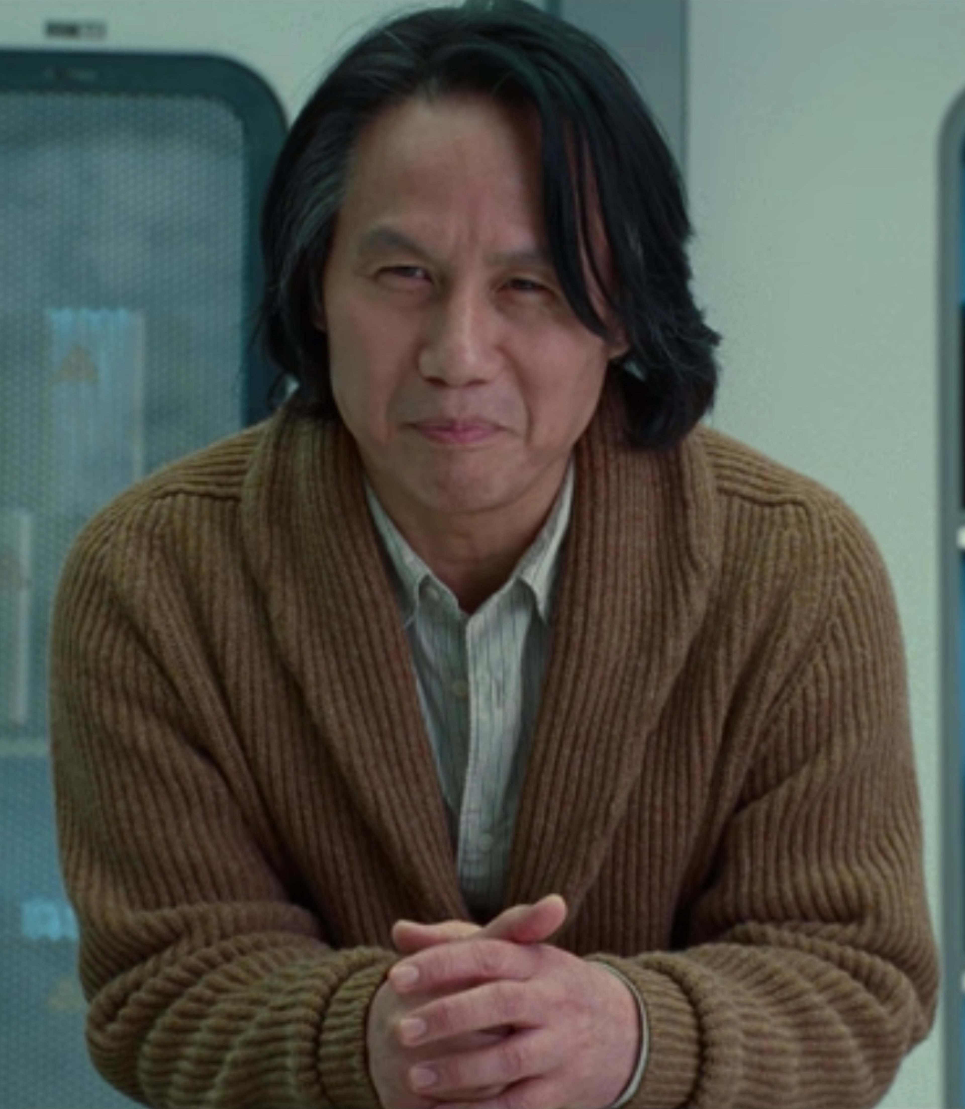 A middle-aged Asian man in a brown sweater sitting in an office or laboratory setting