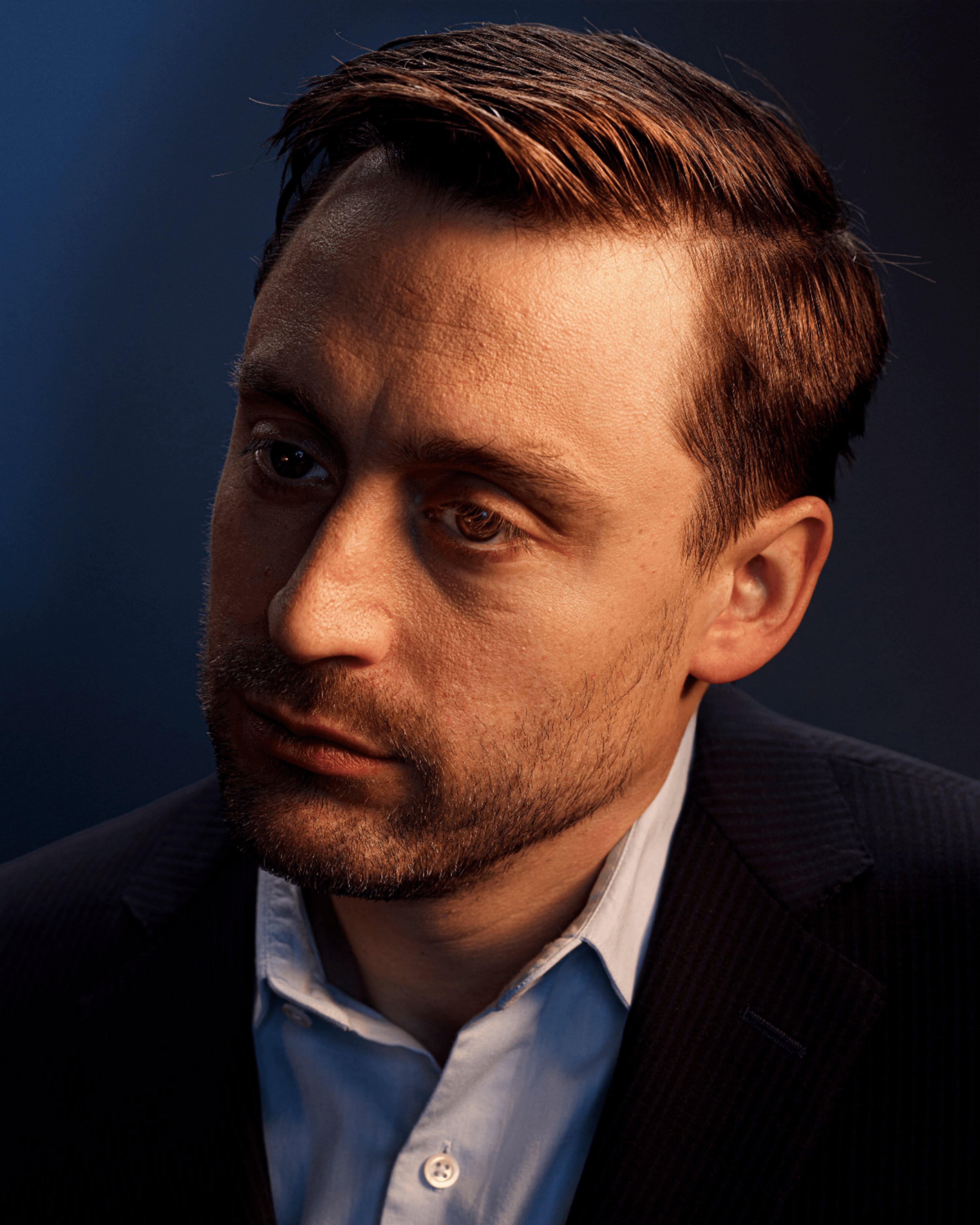 A close-up portrait of a serious-looking man with short brown hair and a beard, wearing a blue collared shirt.