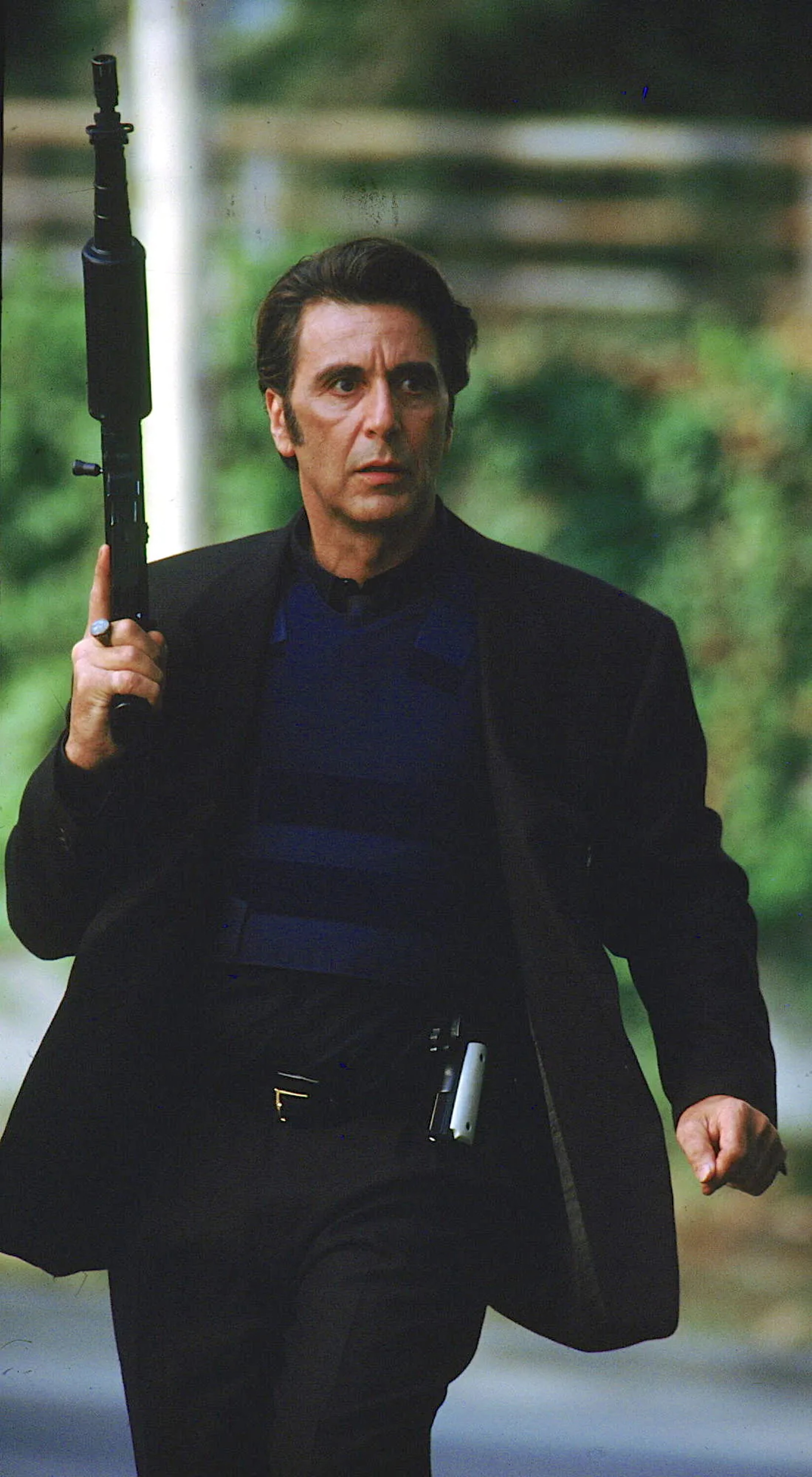 A middle-aged man in a dark suit holding a gun in an outdoor setting