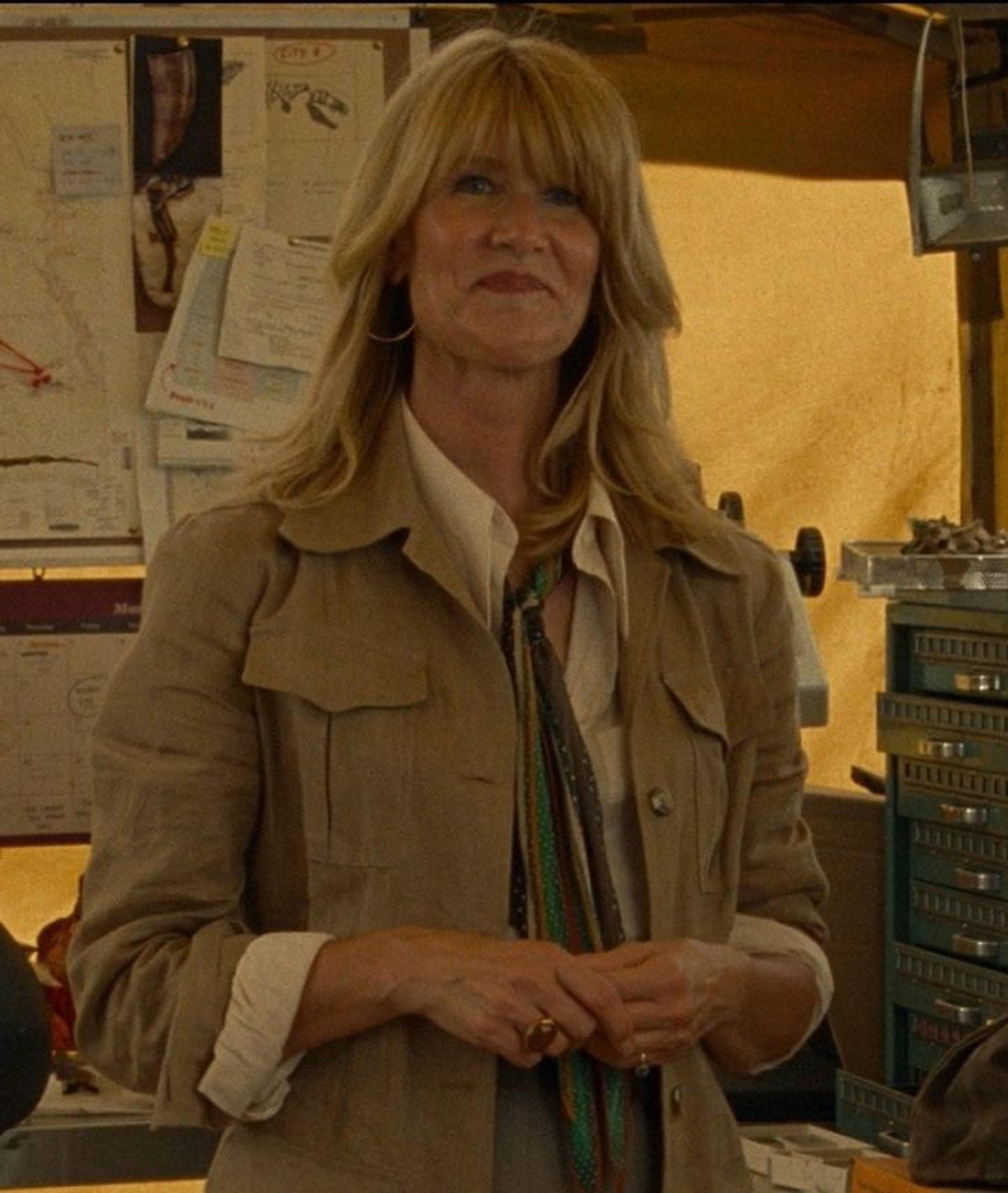 A middle-aged woman with blonde hair wearing a tan trenchcoat standing in a cluttered room