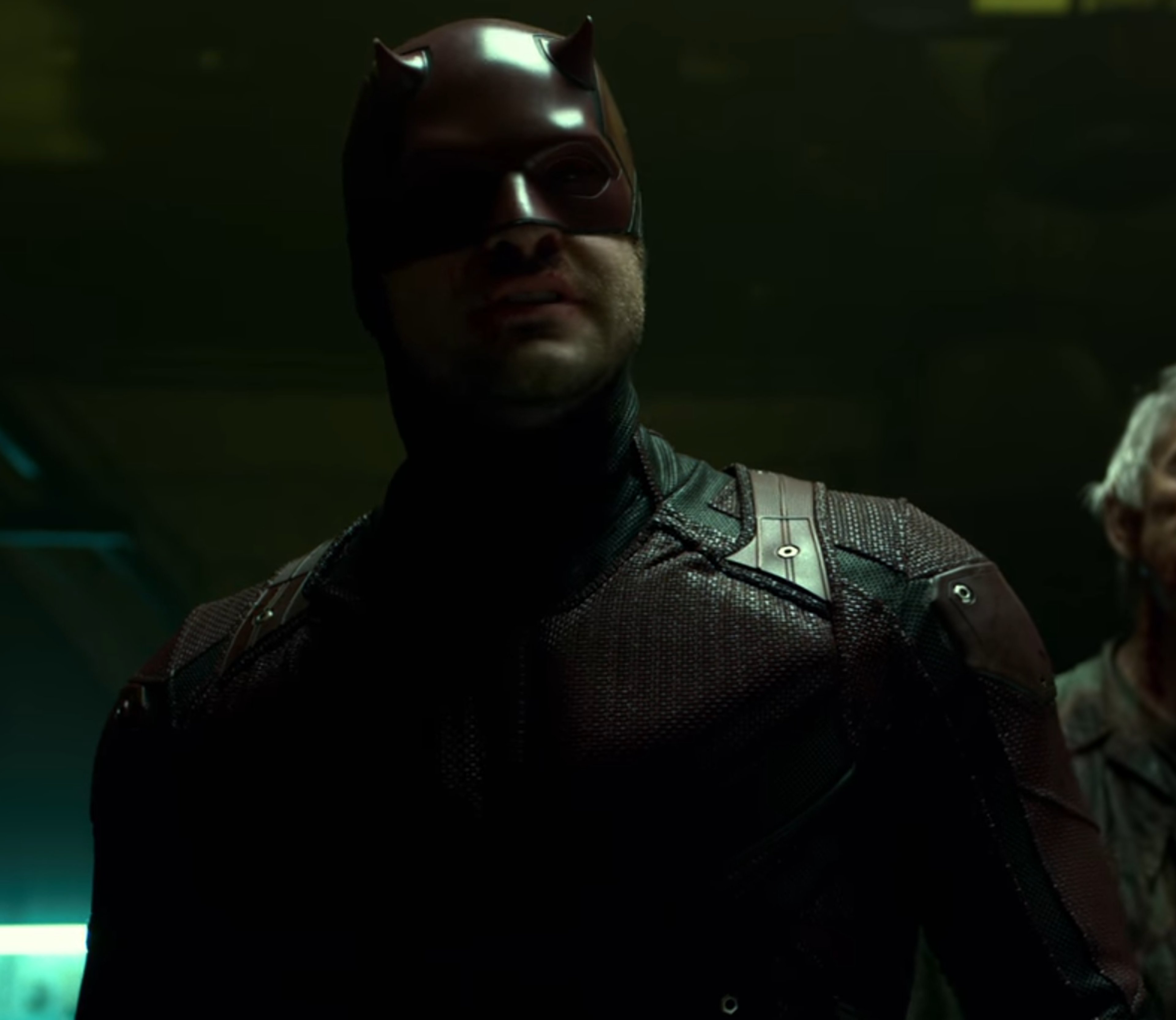 A man in a dark superhero costume with a mask covering the upper half of his face, standing in a dimly lit environment.