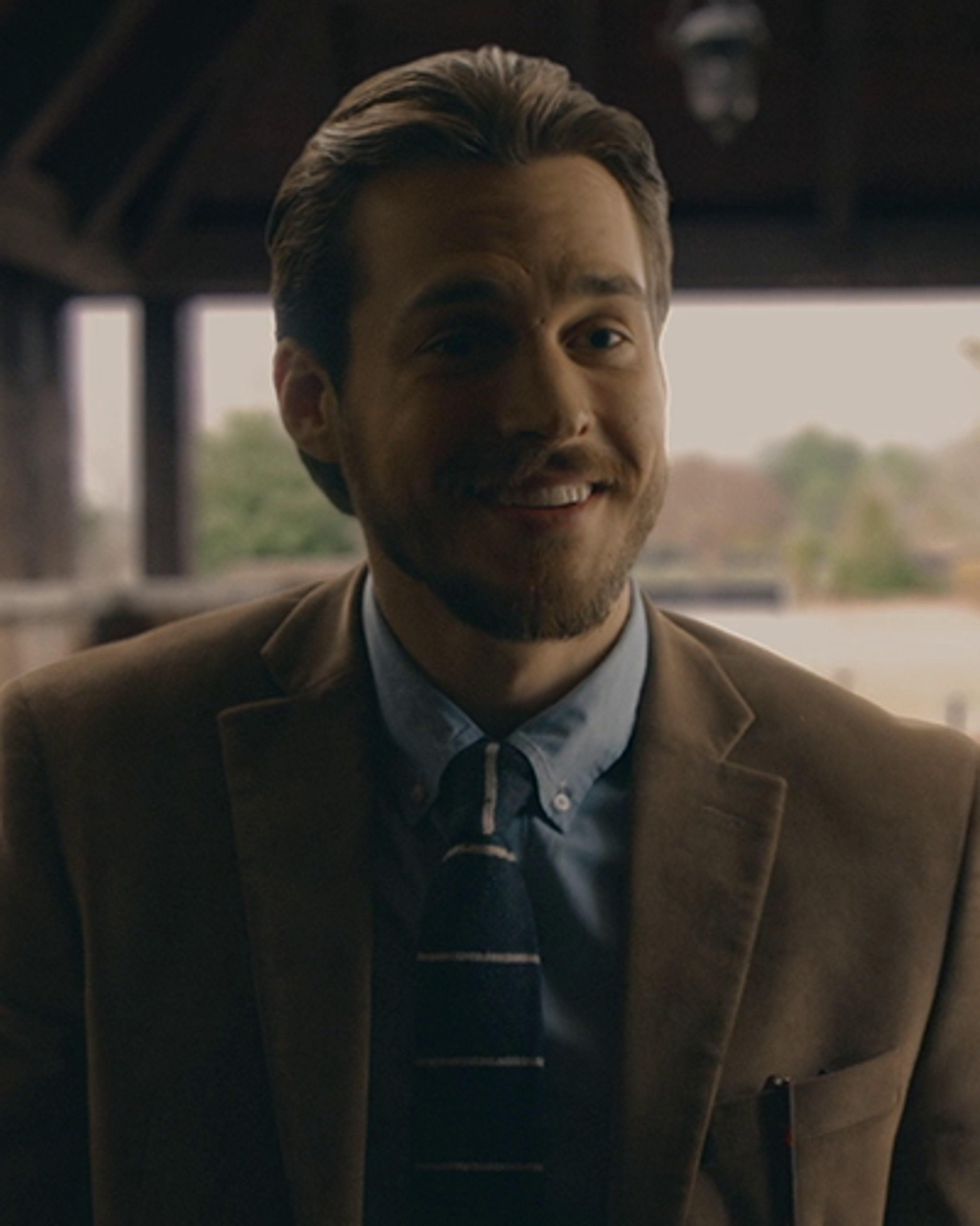 A man with short brown hair and a beard, wearing a suit and tie, with a charming expression on his face.