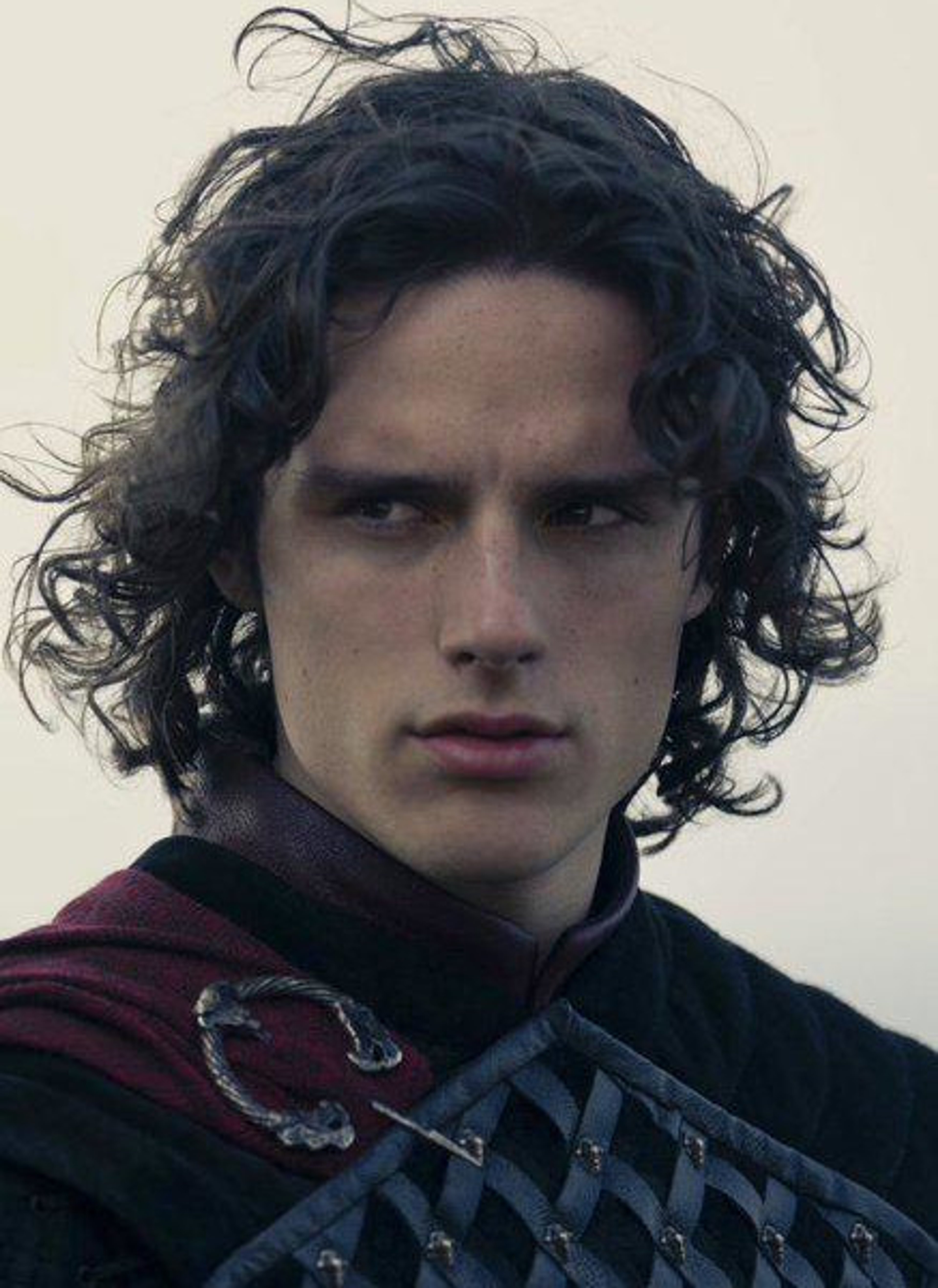 A young man with dark curly hair and a serious expression, wearing a dark fantasy-style outfit.