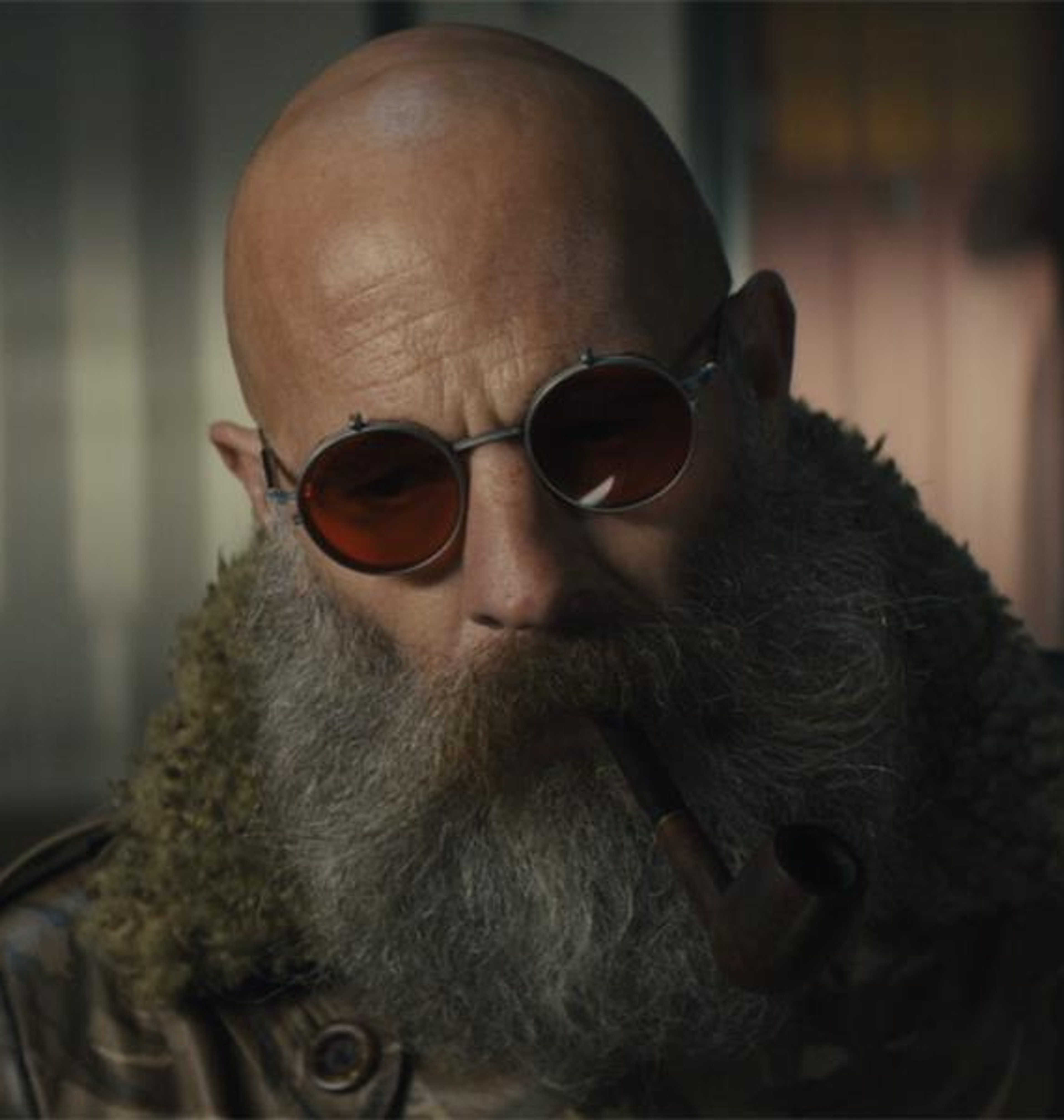 A bald, bearded man with sunglasses wearing a fur-lined coat in a post-apocalyptic setting.
