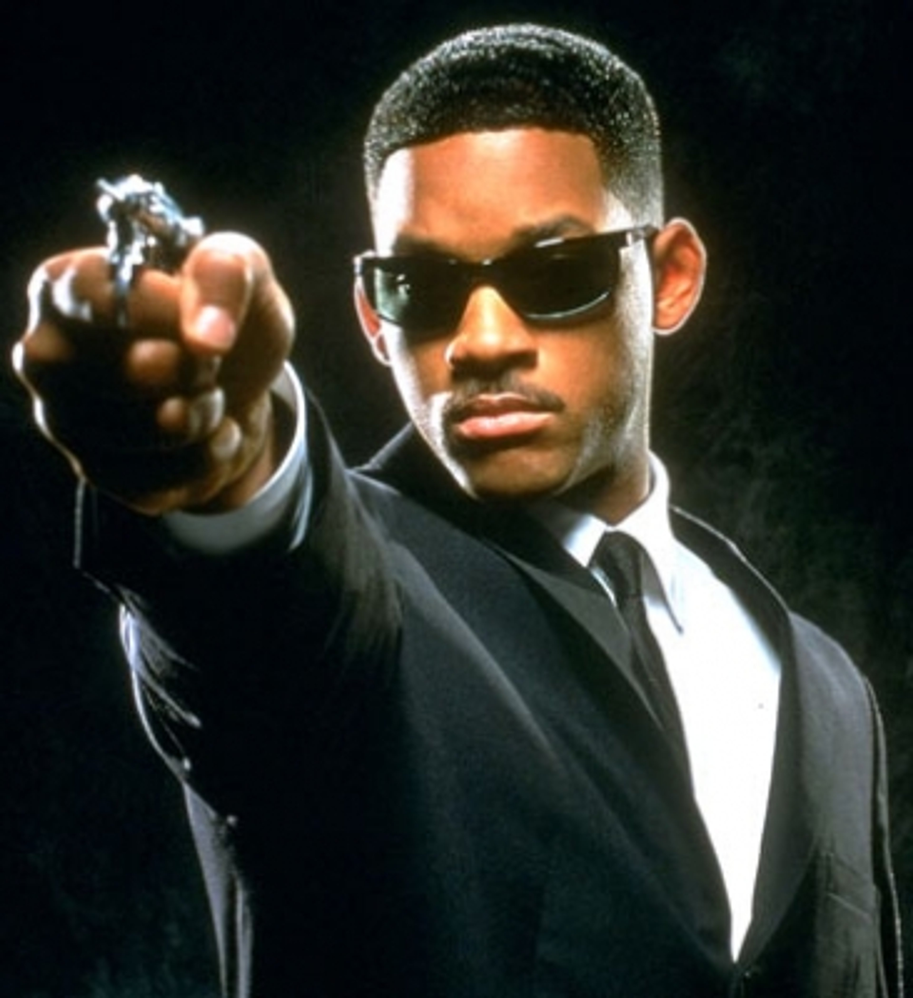 A Black man in a suit holding a gun and looking directly at the camera