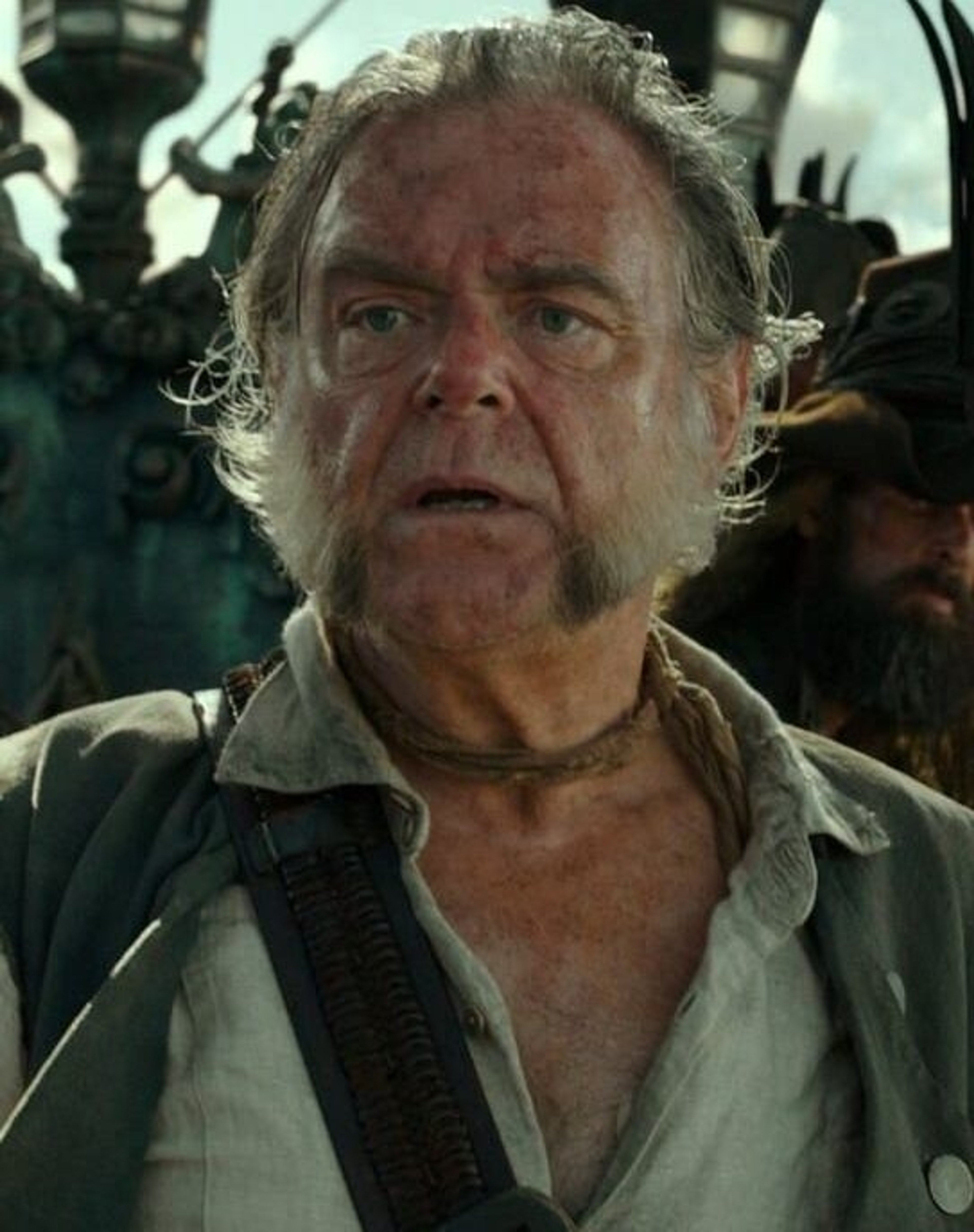 An older man with long, graying hair and a weathered face, wearing a white shirt, dark vest, and bandana.