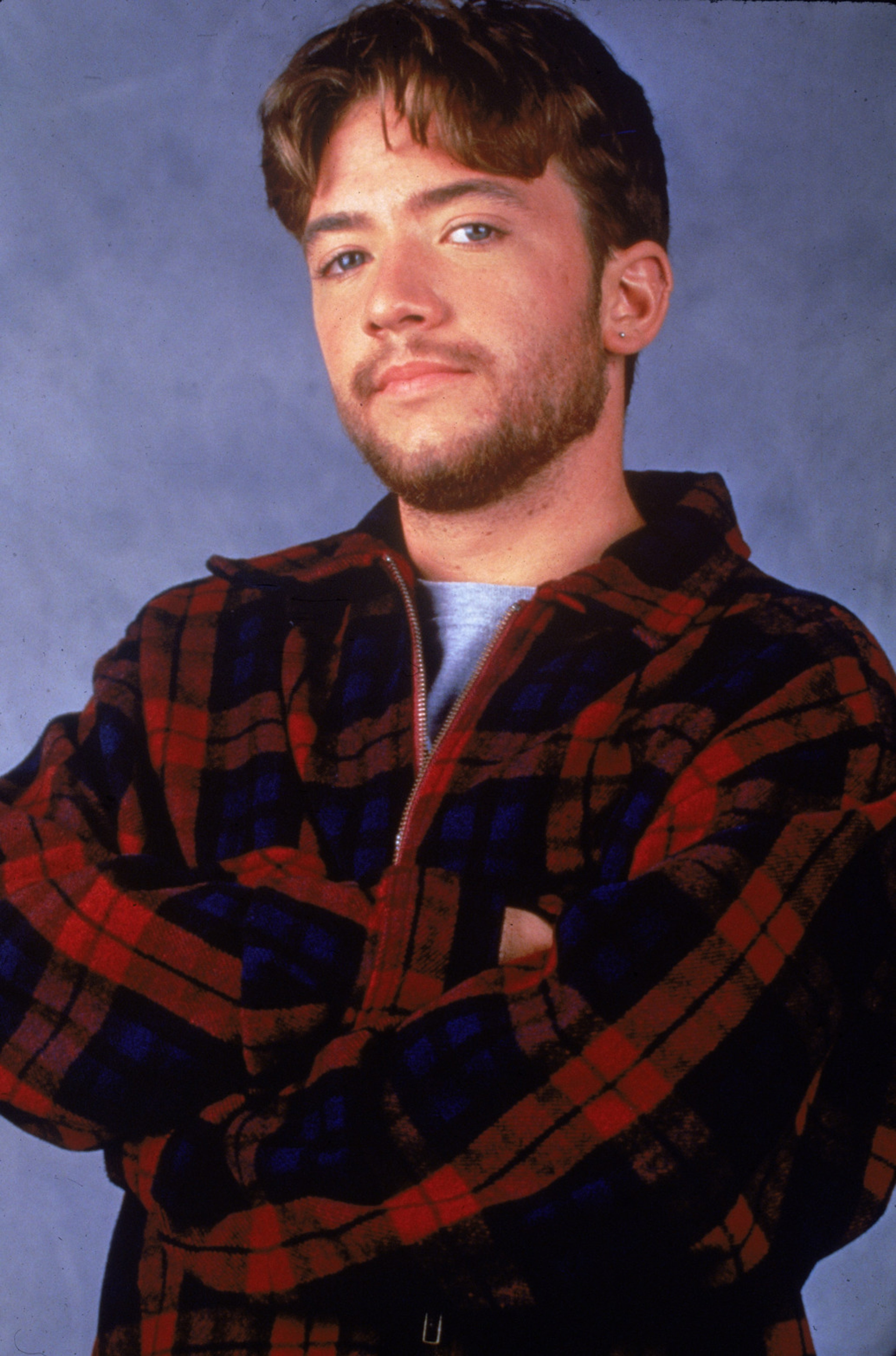 A young man with short dark hair and a beard, wearing a red and black plaid shirt and standing with his arms crossed.