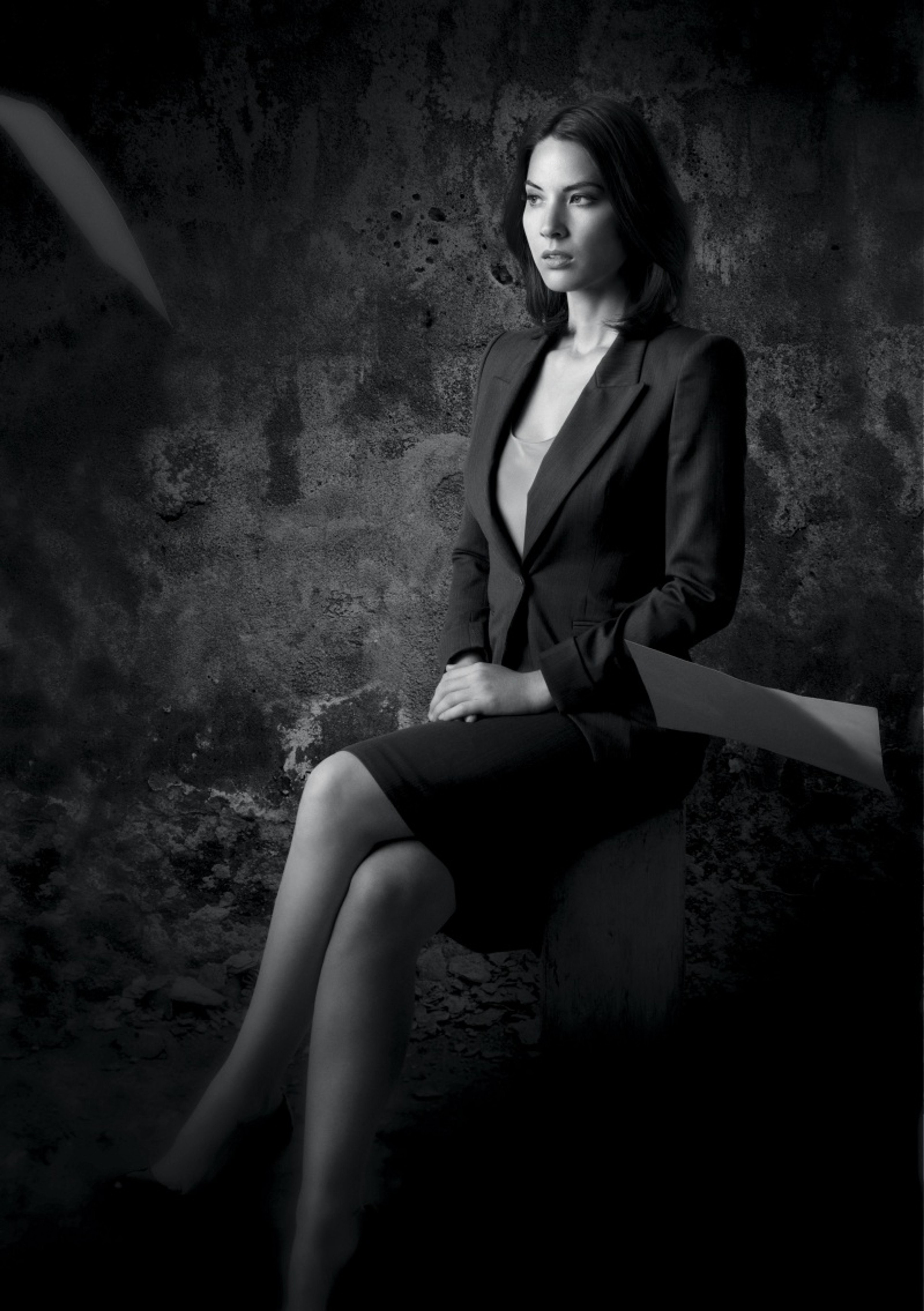 A woman in a black suit jacket sitting on a stone surface with a serious expression