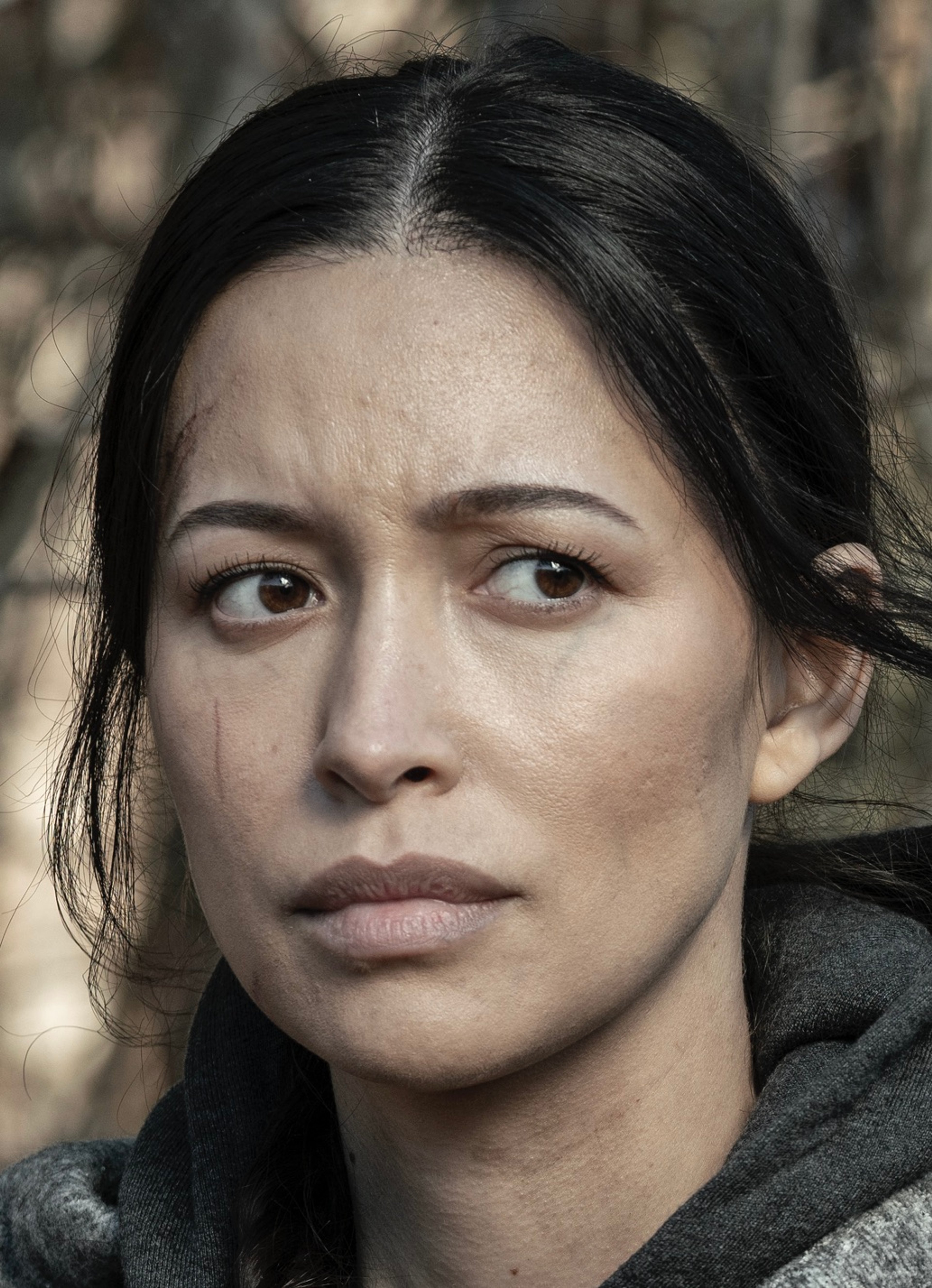 A woman with dark hair and a serious expression, wearing a dark jacket in a post-apocalyptic setting.