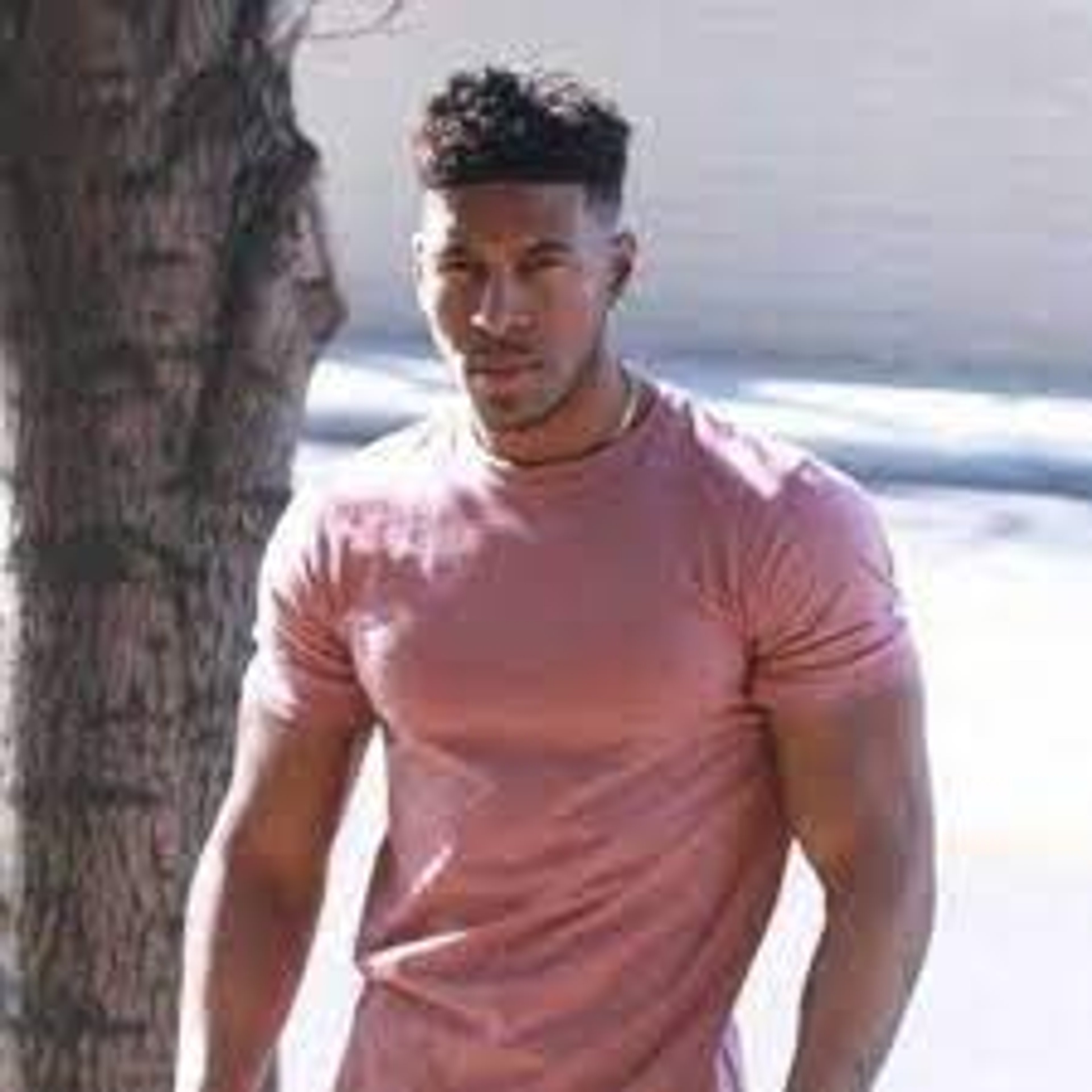 A young, muscular Black man wearing a pink t-shirt standing in front of a body of water